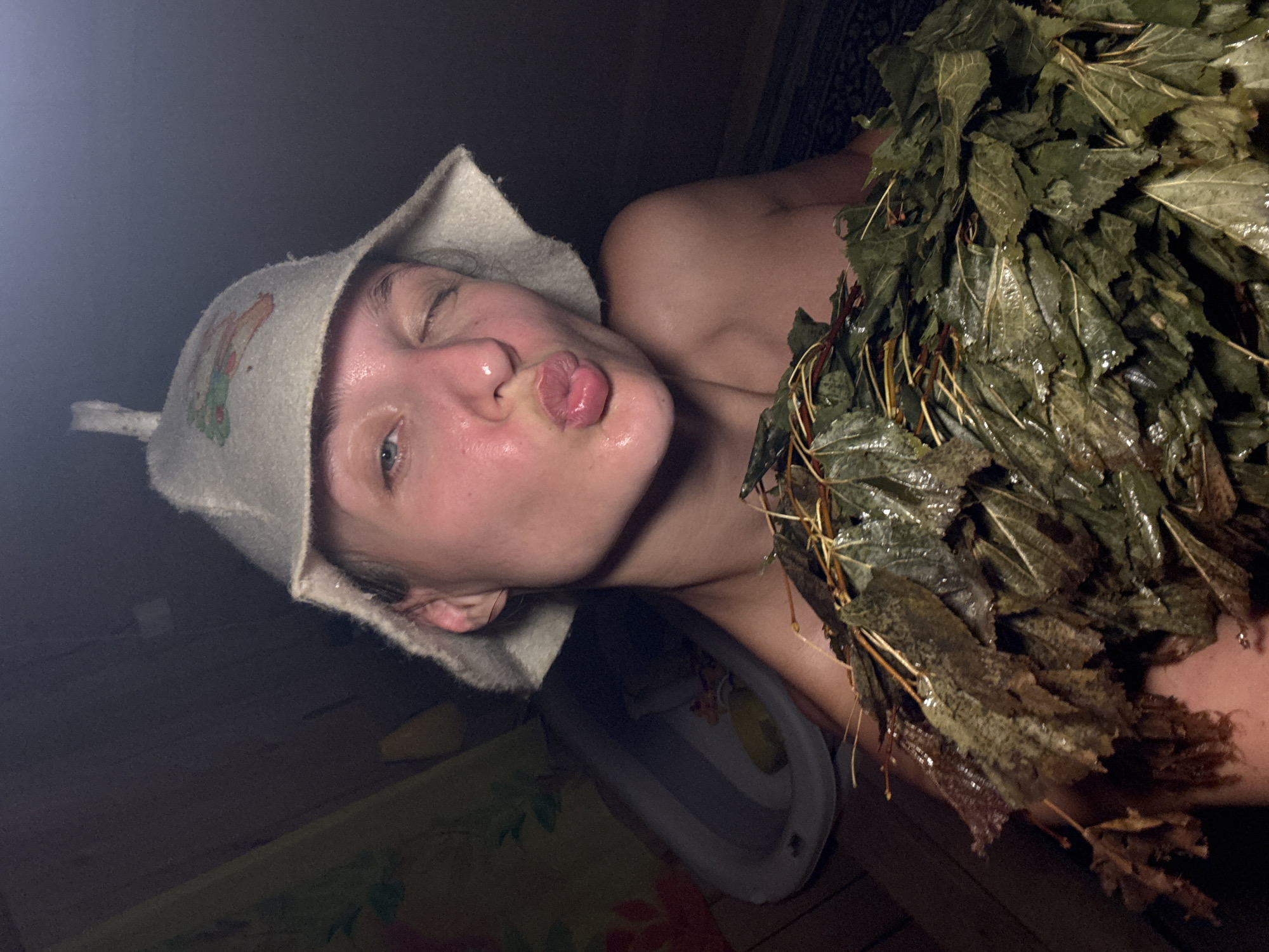 A young woman with wet skin and a light-colored sauna hat adorned with a cartoon-like drawing of an animal is in a dimly lit, rustic sauna. She is winking and puckering her lips towards the camera. She is holding a bundle of wet, green leaves, possibly a venik, in front of her chest. In the background, a grey inflatable bathtub and a patterned curtain are partially visible, suggesting a domestic or traditional sauna setting in Sharkan, Russia. The lighting is low, creating a warm and steamy atmosphere.