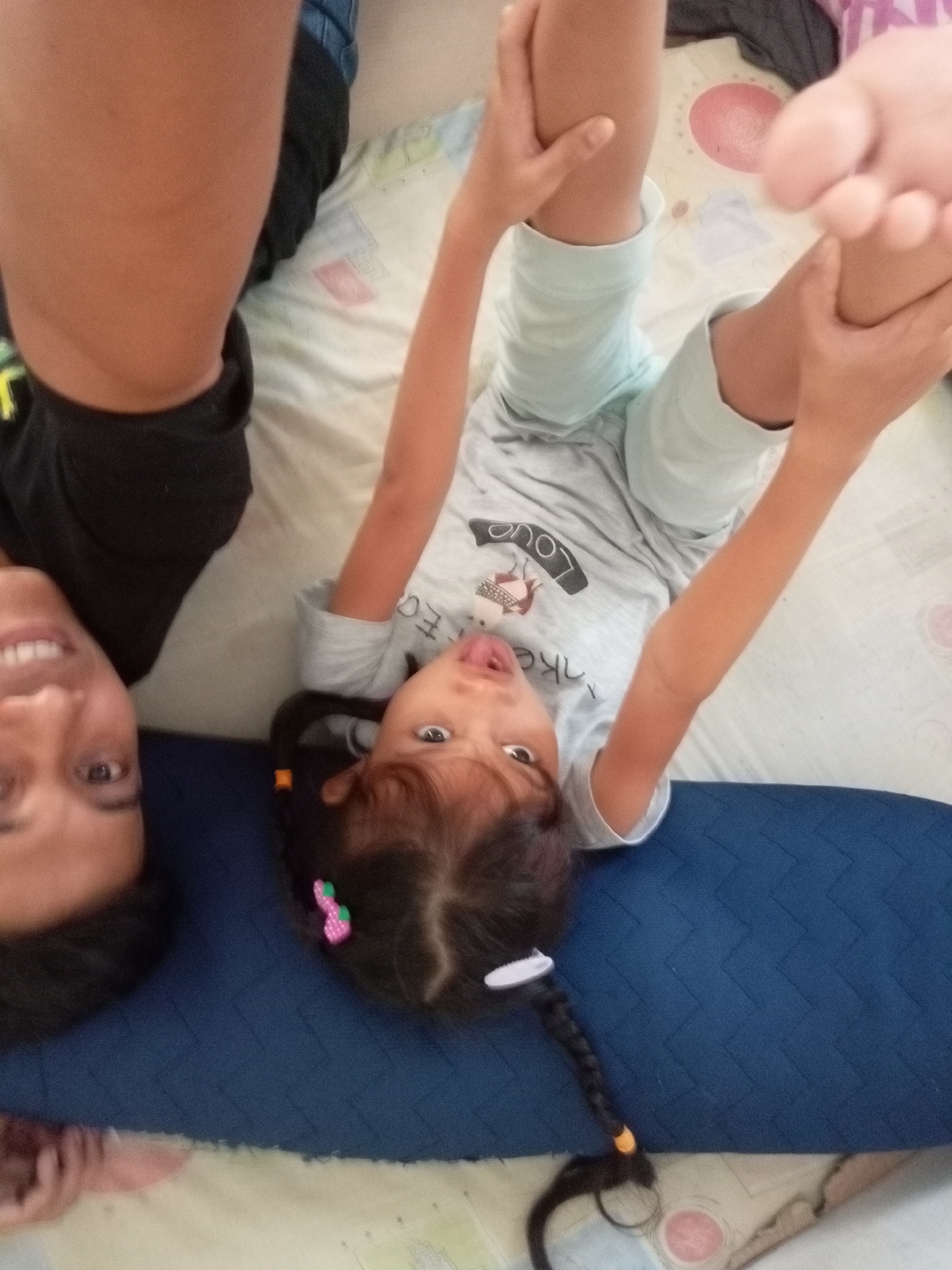 The image depicts two individuals, an adult and a child, lying on their backs on a blue mat with a patterned bedspread beneath them. The adult, positioned on the left, is looking upwards with a slight smile, their head partially visible. The child, to the right, is lying with their legs in the air, wearing a light-colored t-shirt and shorts. The child has dark hair styled in a braid and is sticking their tongue out. Both individuals appear to be indoors. The image is oriented such that the heads are towards the bottom and the feet towards the top.