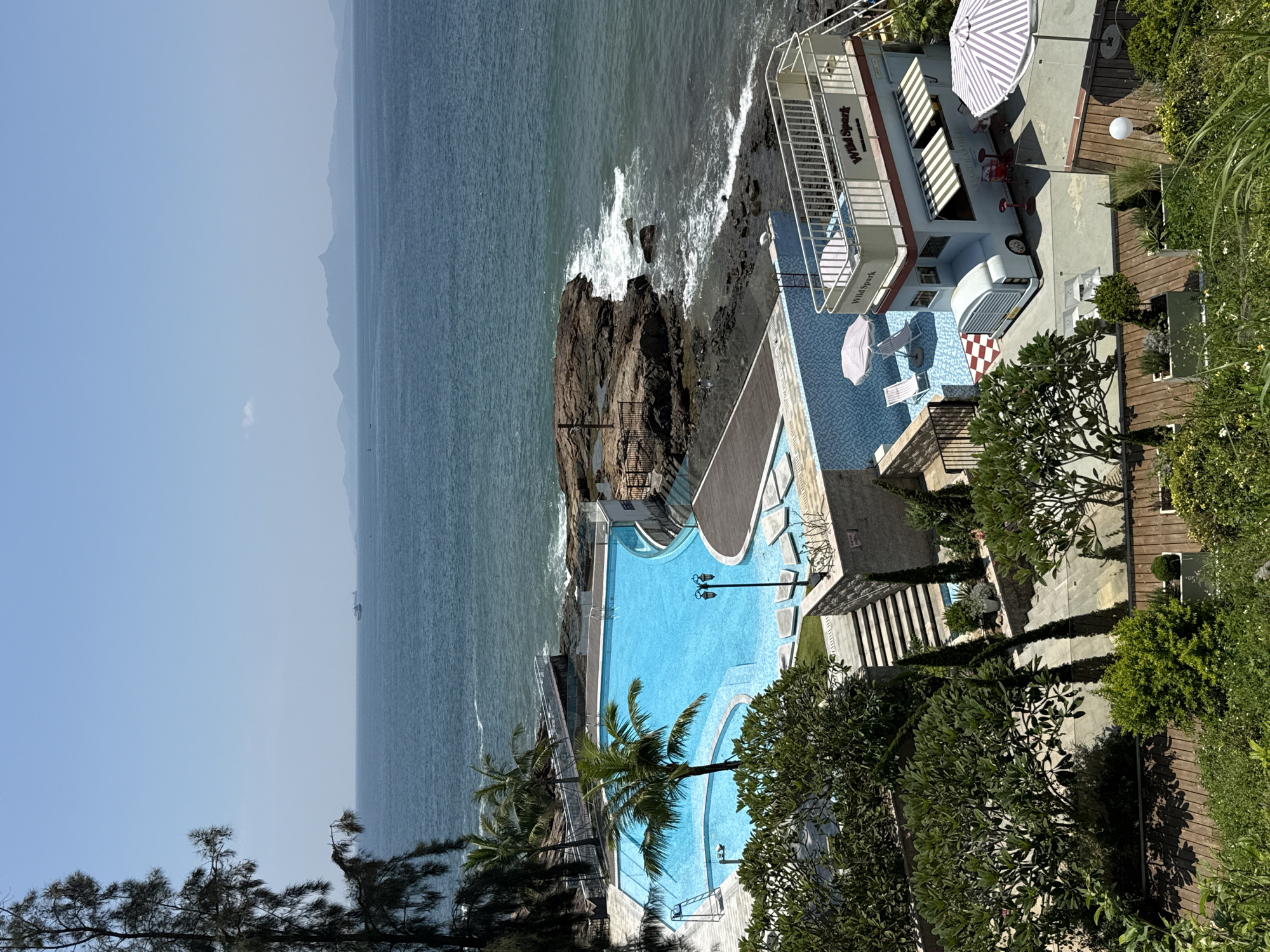 This is an eye-level, medium shot of a resort or hotel swimming pool area located in Longgang District, China, with the ocean in the background. The swimming pool is a bright turquoise color and is surrounded by a paved area with lounge chairs and umbrellas. To the right of the pool is a small, white and blue double-decker bus-like structure with an awning and seating area. Beyond the pool, a rocky coastline meets the blue ocean, and in the distance, there are mountains and ships visible on the horizon. Lush green trees and plants are visible in the foreground, suggesting a tropical or subtropical climate. The sky is clear and blue. There are no people visible in the image.