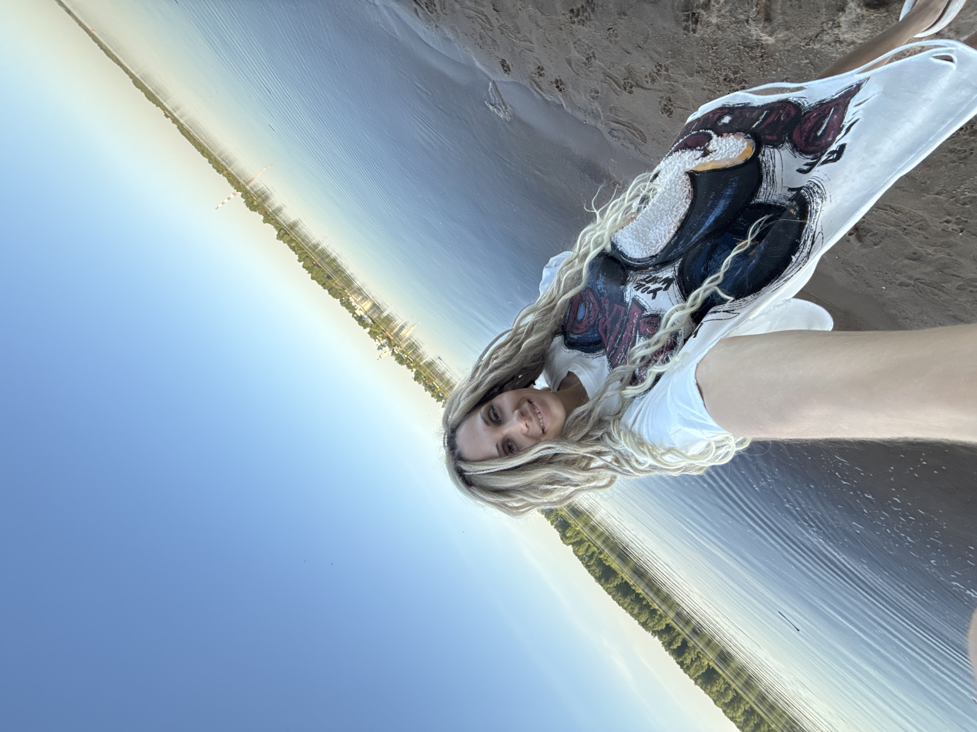 A woman with long blonde dreadlocks smiles at the camera, wearing a white t-shirt with a dark graphic. She stands on a sandy shore next to a body of water, likely a river or lake. Across the water, a line of trees runs along the bank. In the distance, a city skyline is visible. The location is likely Rusanovo, Russia.