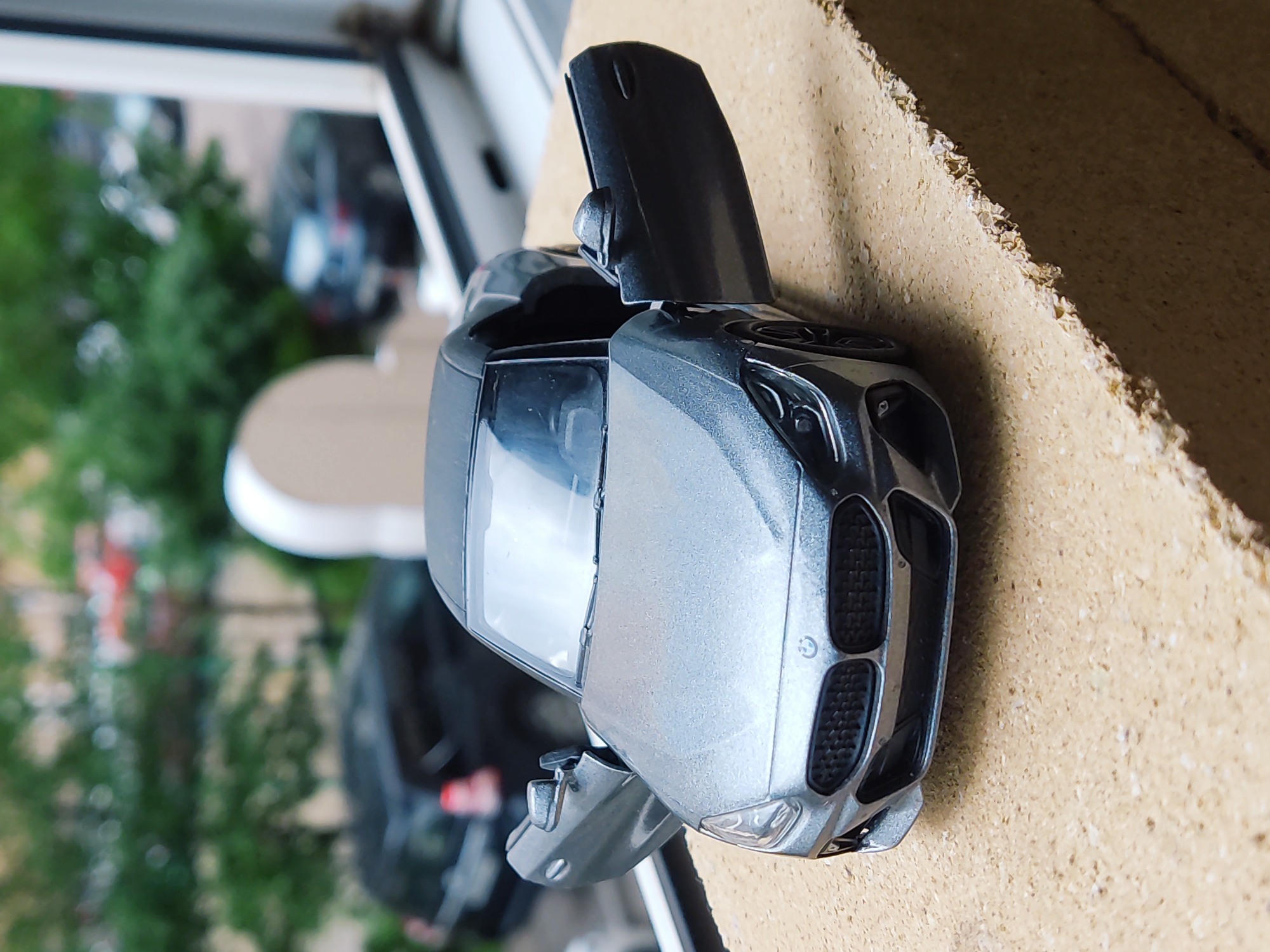 A gray BMW toy car with open doors sits on a rough, beige surface. The background shows a window with greenery visible outside, suggesting a view of trees and possibly other buildings. The scene appears to be indoors, likely on a windowsill or ledge. There are no people visible in the image. The photograph is taken at an angle, emphasizing the front of the toy car. There is no indication of the city of Krasnodar, Russia.