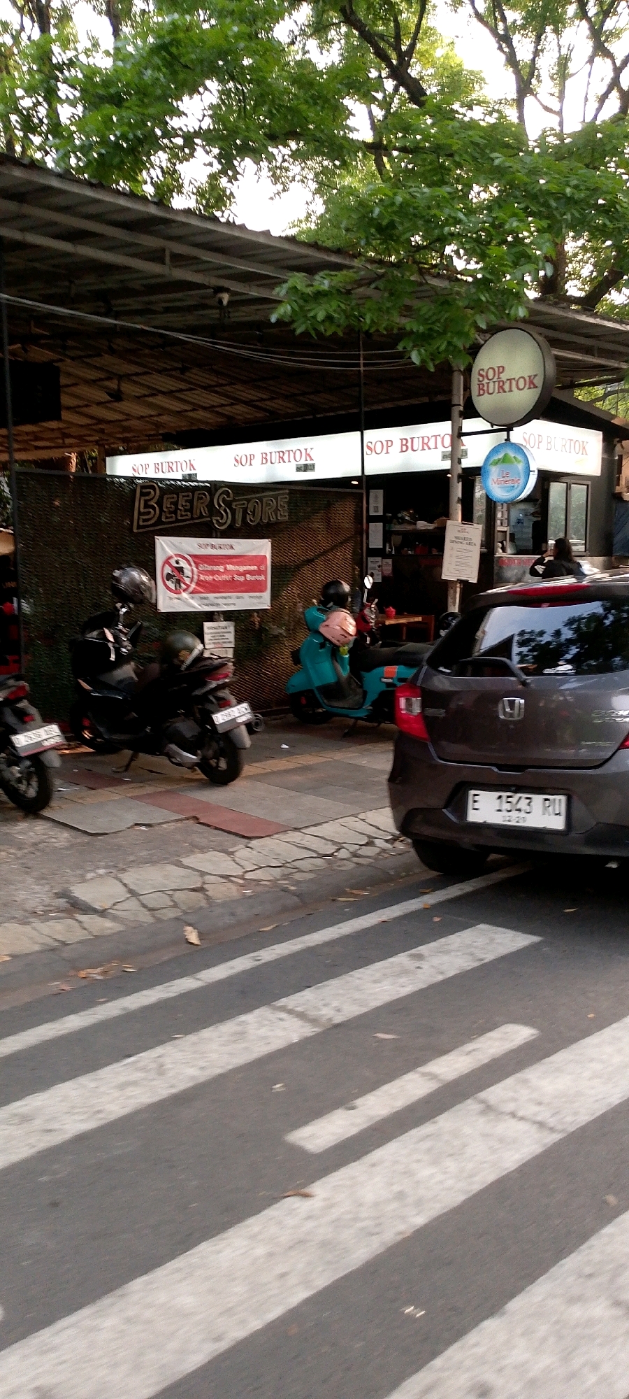 The image depicts a street scene in Bandung City, Indonesia, featuring a storefront with prominent signage and a crosswalk in the foreground. The storefront displays the words "Beer Store" in large, stylized letters, along with smaller signs that read "SOP BURTOK." A circular sign above the storefront also reads "SOP BURTOK." Several motorcycles are parked in front of the store, and a dark-colored car is visible on the right side of the image, with its license plate reading "E 1543 RU." A pedestrian crosswalk with white stripes is painted on the asphalt in the foreground. Trees with dense green foliage overhang the scene from above. The overall impression is of a commercial area with food and drink establishments.