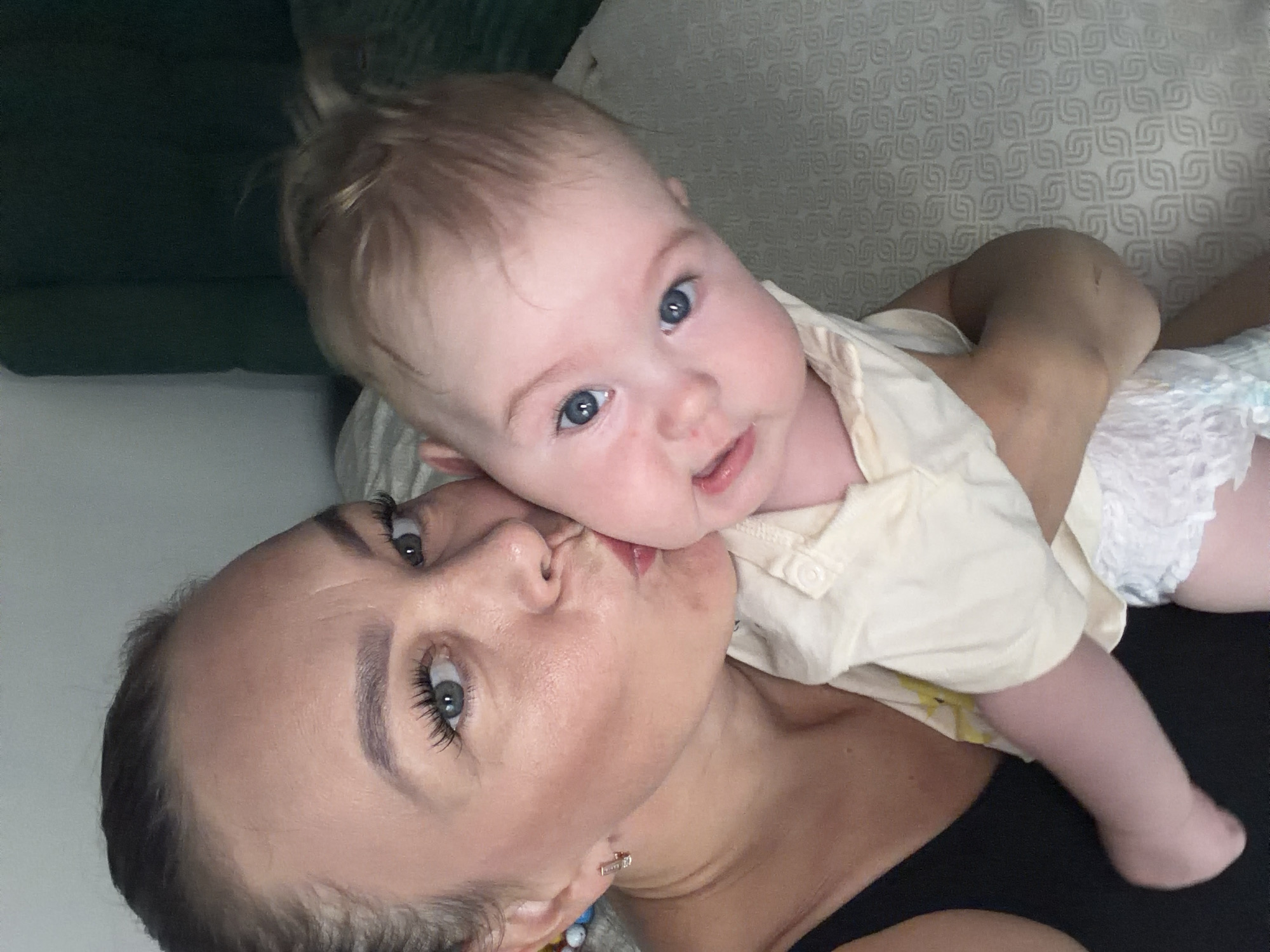 A woman is kissing a baby on the cheek in a close-up selfie. The woman has dark hair pulled back, blue eyes, and is wearing a black tank top and a small hoop earring. The baby has blonde hair and blue eyes and is wearing a light-colored shirt and a diaper. They are indoors, on a surface that appears to be a bed with a patterned textured sheet. The background is blurred, with a dark green object that might be a headboard or furniture visible behind them. The scene is lit by an overhead light source.