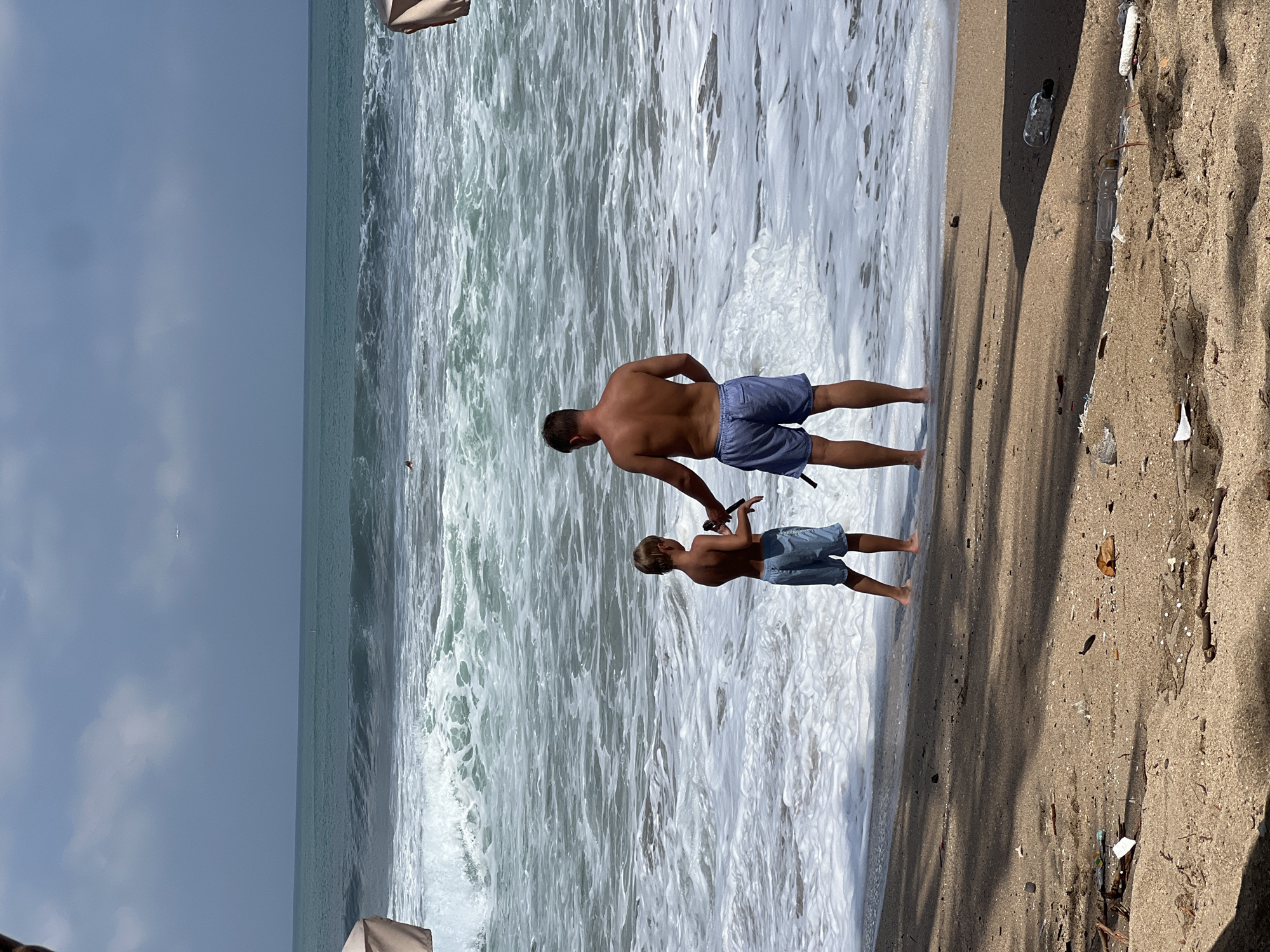 The image shows a man and a boy on a beach in Kuta, Indonesia. The man is wearing blue shorts and the boy is also wearing blue shorts. They are standing near the water's edge with the waves crashing around them. The man appears to be holding a stick or a fishing rod, and the boy is facing him, possibly interacting with the stick. The beach is sandy with some debris visible, including plastic bottles and sticks. The sky is partly cloudy and the sun appears to be shining, suggesting it is daytime. The overall scene depicts a casual day at the beach, with the father and son enjoying the ocean.