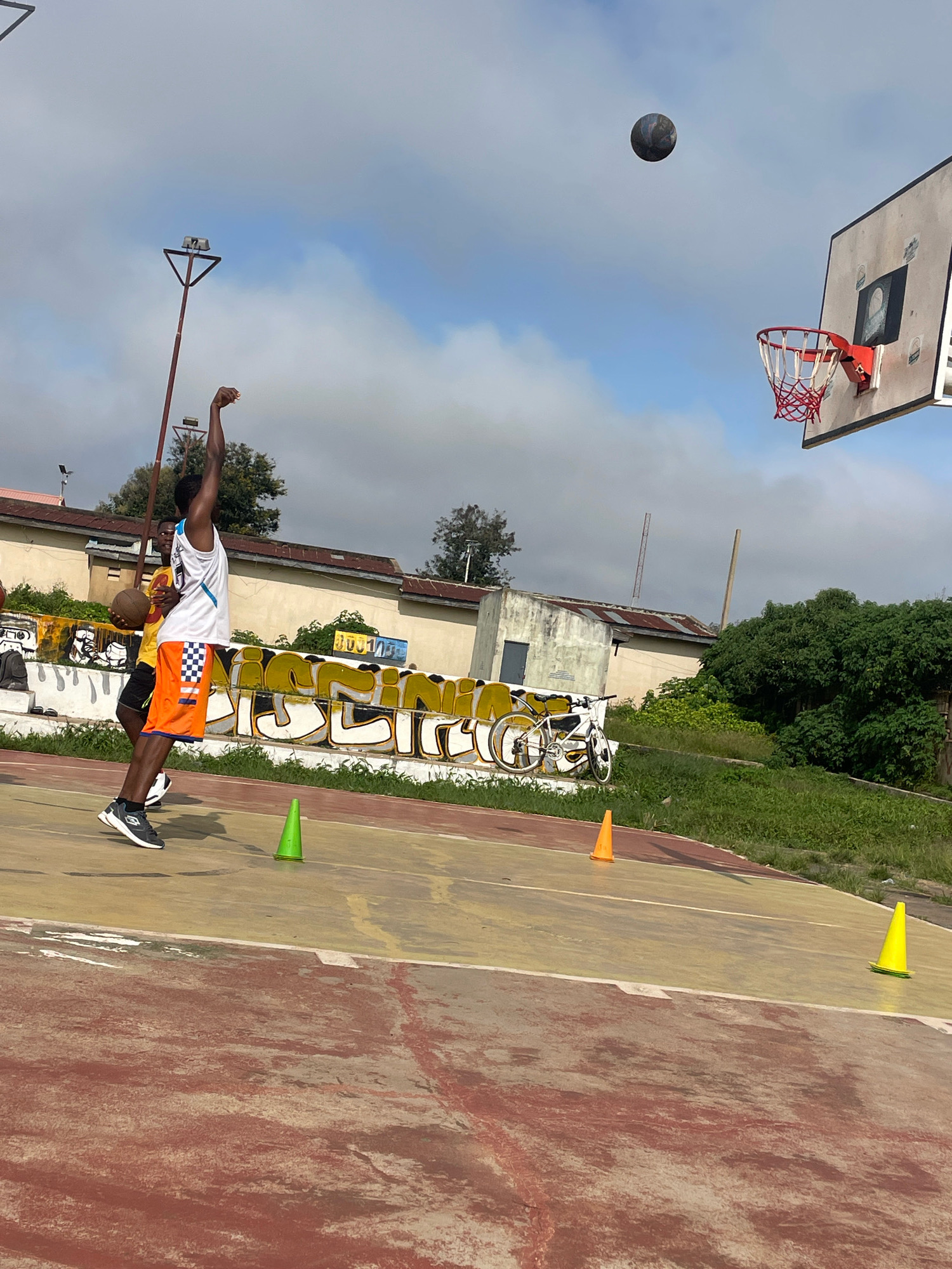 In Gura Top, Nigeria, on an outdoor basketball court, two young men are engaged in a basketball practice session. The player in the foreground, wearing a white tank top and orange and white checkered shorts, is in the motion of shooting a basketball. His arm is extended upwards, and the ball is airborne above him, heading towards the basket. Another player, partially visible behind him and to his left, is holding a basketball. The court surface is a mix of red and yellow concrete, marked with lines. Green and orange traffic cones are placed on the court, likely for drills. In the background, there is a graffiti-covered wall with a white bicycle leaning against it. Beyond the wall, buildings with corrugated metal roofs and trees are visible under a cloudy sky. The scene suggests a casual, recreational sports activity taking place in an urban or semi-urban setting.