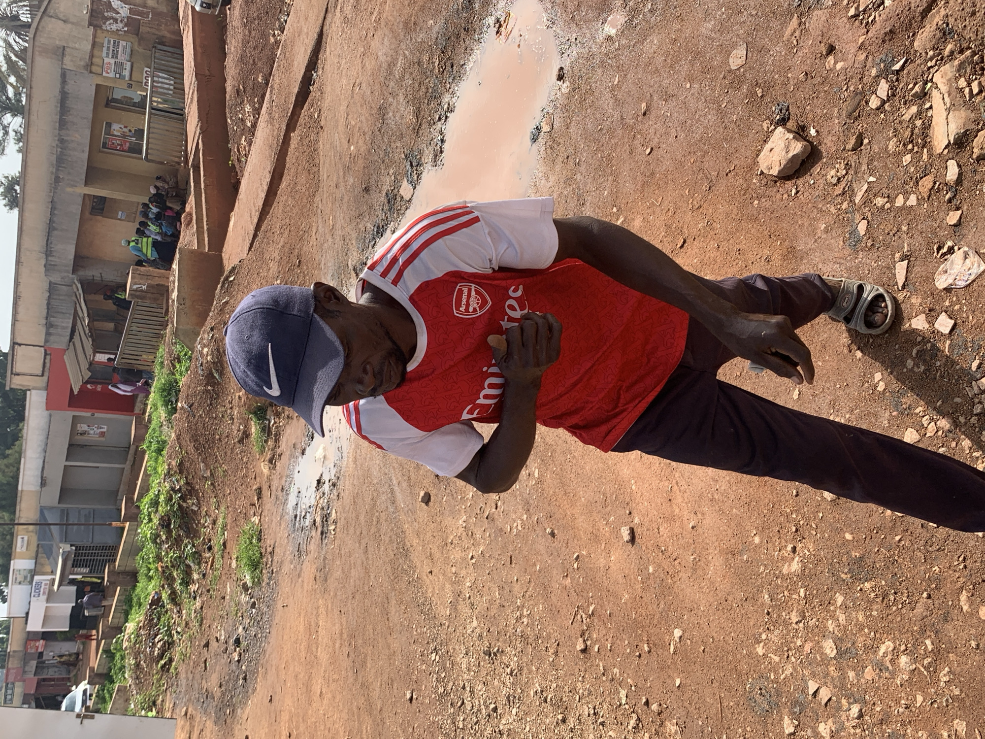 In Gura Top, Nigeria, a man wearing a blue baseball cap and a red Arsenal soccer jersey with white sleeves stands on a dirt road. The jersey has the "Emirates" logo and an Arsenal crest. He is also wearing dark pants and gray sandals. His right hand is raised, giving a thumbs-up gesture. The ground is reddish-brown dirt with scattered small stones and debris. To the right and slightly behind the man, a large puddle of muddy water is visible. In the background, there are low-rise buildings with storefronts and people gathered in front of one building. Vegetation and more debris are present in the mid-ground, between the man and the buildings. The scene appears to be an outdoor market or commercial area.