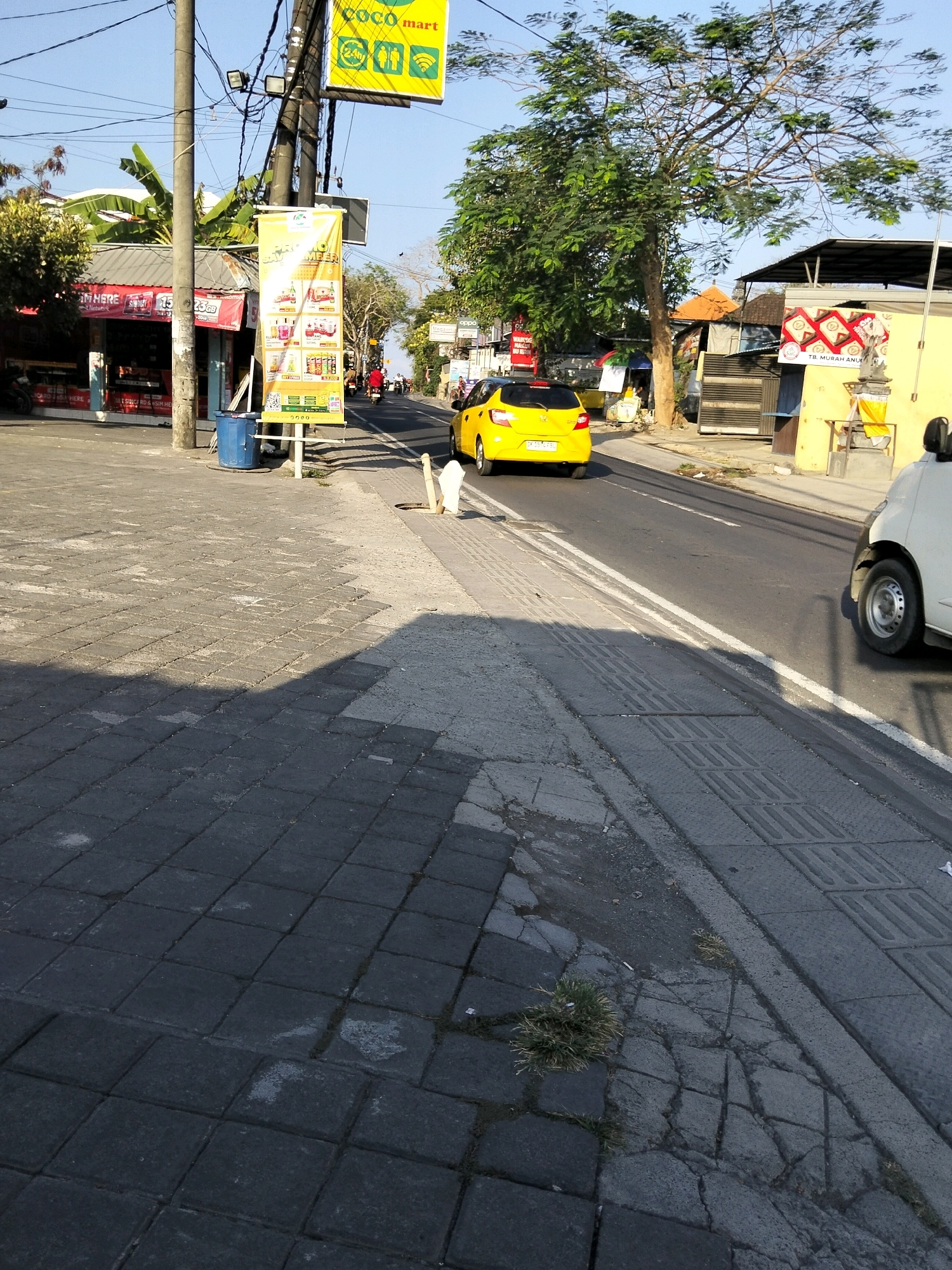 The image captures a street scene in Kuta Selatan, Indonesia, during daylight hours under a clear blue sky. The foreground is dominated by a gray tiled sidewalk, some sections of which appear cracked and uneven, with small patches of grass growing through the gaps. A tall, black utility pole stands on the left side of the sidewalk, supporting multiple wires and lights. A large blue trash bin is situated at the base of the pole.
To the left of the sidewalk, a small shop with a red and white sign is visible, displaying what appear to be product advertisements. Behind the shop, banana leaves and other foliage suggest a tropical environment. A yellow banner with text and images of products hangs from the utility pole, advertising what seems to be a local mart.
The street itself is paved asphalt, with distinct white lane markings. A bright yellow car is in motion on the right side of the road, heading away from the viewer. A white vehicle is partially visible on the far right of the image, also on the road. Further down the street, other cars and motorbikes can be seen, along with more buildings and trees. The overall impression is of a busy street in a commercial area, typical of a town or city in Indonesia.