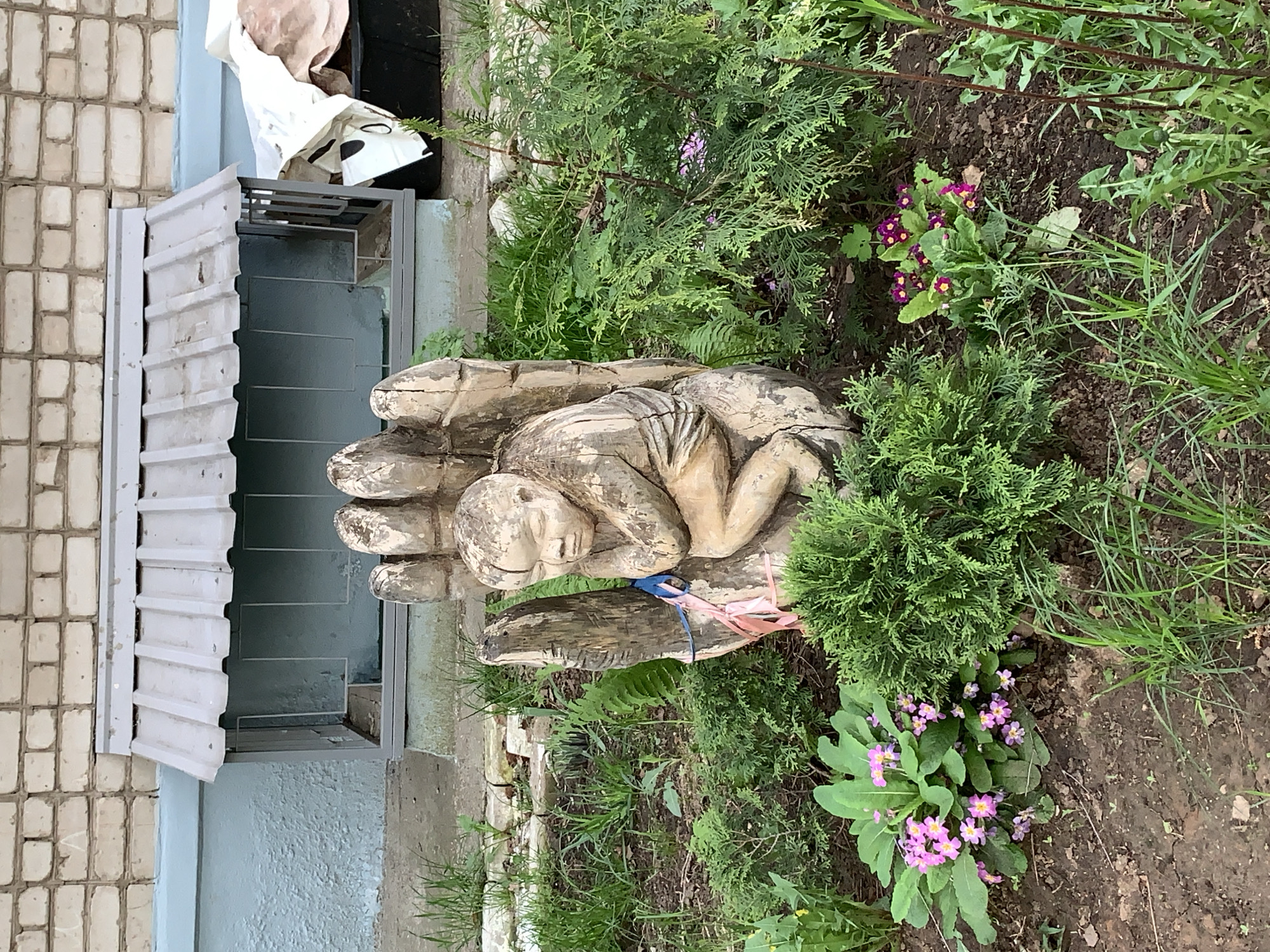 The image depicts a wooden sculpture of a person nestled in a large hand, set amidst greenery and flowers, likely in a garden setting. The sculpture appears to be the focal point, surrounded by small evergreen bushes, grass, and blooming primroses with pink flowers. The background includes a building with a partially open window and a brick wall. Given the prompt, the location is presumed to be in the city of Chaikovsky, Russia, though no specific landmarks are visible in the image to confirm this. The overall scene suggests a peaceful outdoor space with artistic and natural elements.