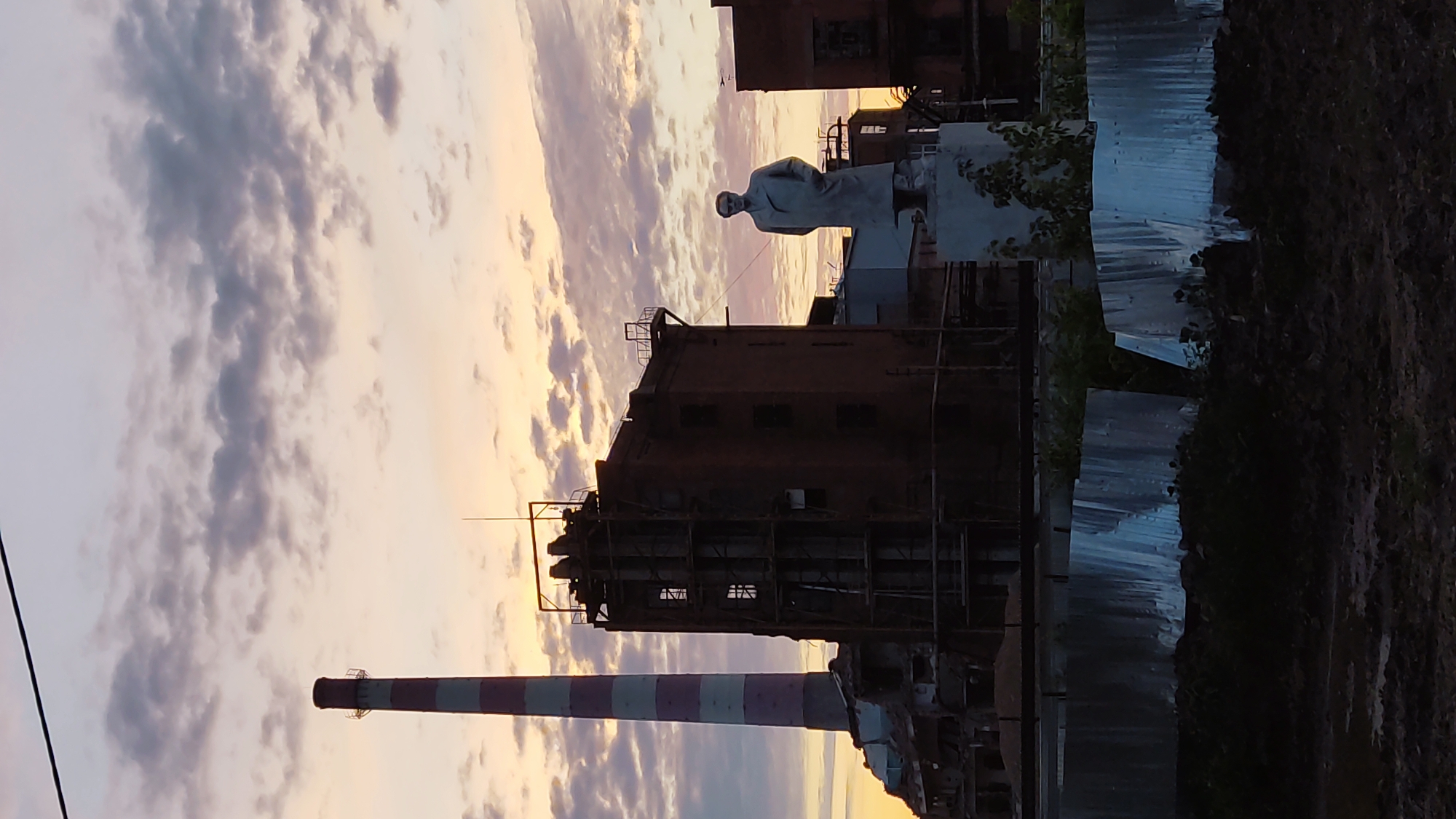 An outdoor vertical, low-angle shot captures an industrial scene at dusk. In the foreground, a corrugated metal fence runs horizontally, partially obscured by dark soil and vegetation. Behind the fence, a statue of a man in a long coat stands on a pedestal. To the left of the statue, a dilapidated brick building with scaffolding is visible. Further left, a tall, red and white striped smokestack rises into the cloudy sky. The sky itself is a gradient of pale orange and pink, with scattered grey clouds. The overall impression is one of an abandoned or disused industrial area bathed in the soft light of sunset. The specific location in Krasnodar, Russia, is not discernible from the visual information provided in the image.