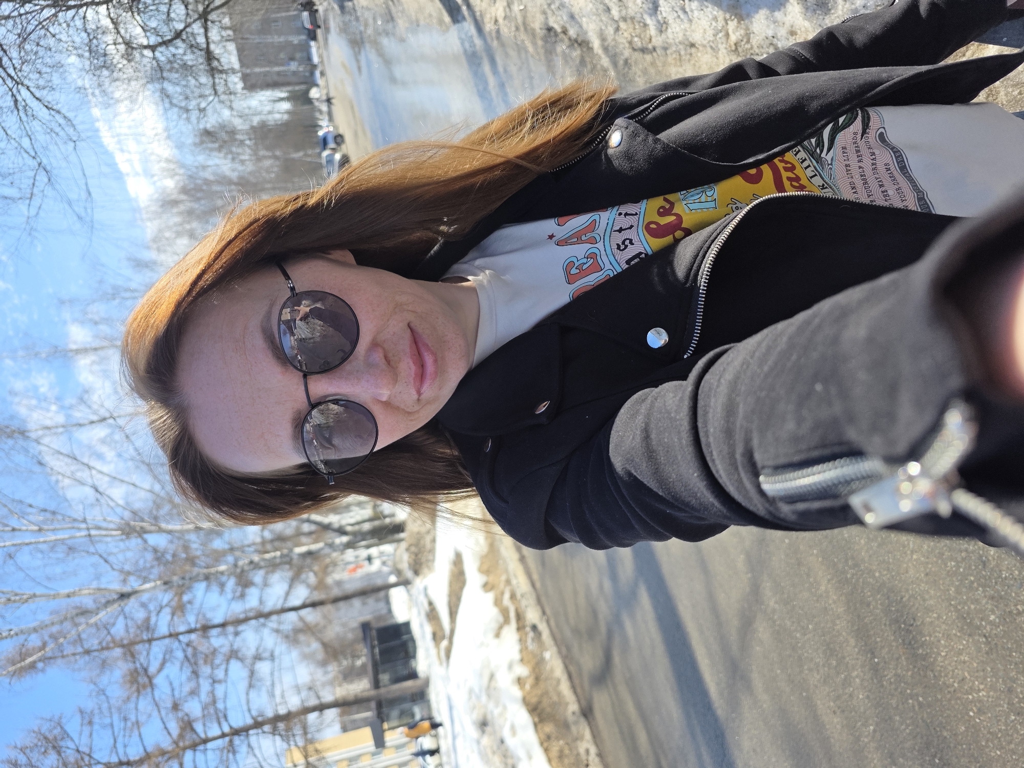 The image presents a selfie of a woman taken outdoors in Chaikovsky, Russia, under clear blue skies typical of late winter or early spring. The subject, a woman with reddish-brown hair and visible freckles on her nose and cheeks, is positioned in the foreground, extending her right arm to hold the camera. She wears dark, round sunglasses, a black jacket with prominent silver zippers and snaps, and a light-colored t-shirt with a colorful graphic design featuring text. Her lips are slightly parted in a faint smile.
The background shows an asphalt road, partially wet and bordered by melting snow and patches of exposed ground. Numerous bare deciduous trees line both sides of the street. In the mid-ground and background, several vehicles are visible on the road, and multi-story buildings can be seen, including a light-colored structure to the left and a grey building further down on the right. Distant figures, including one person in a bright yellow coat, are visible walking on the far left side of the street. The overall environment is illuminated by bright sunlight, casting shadows.