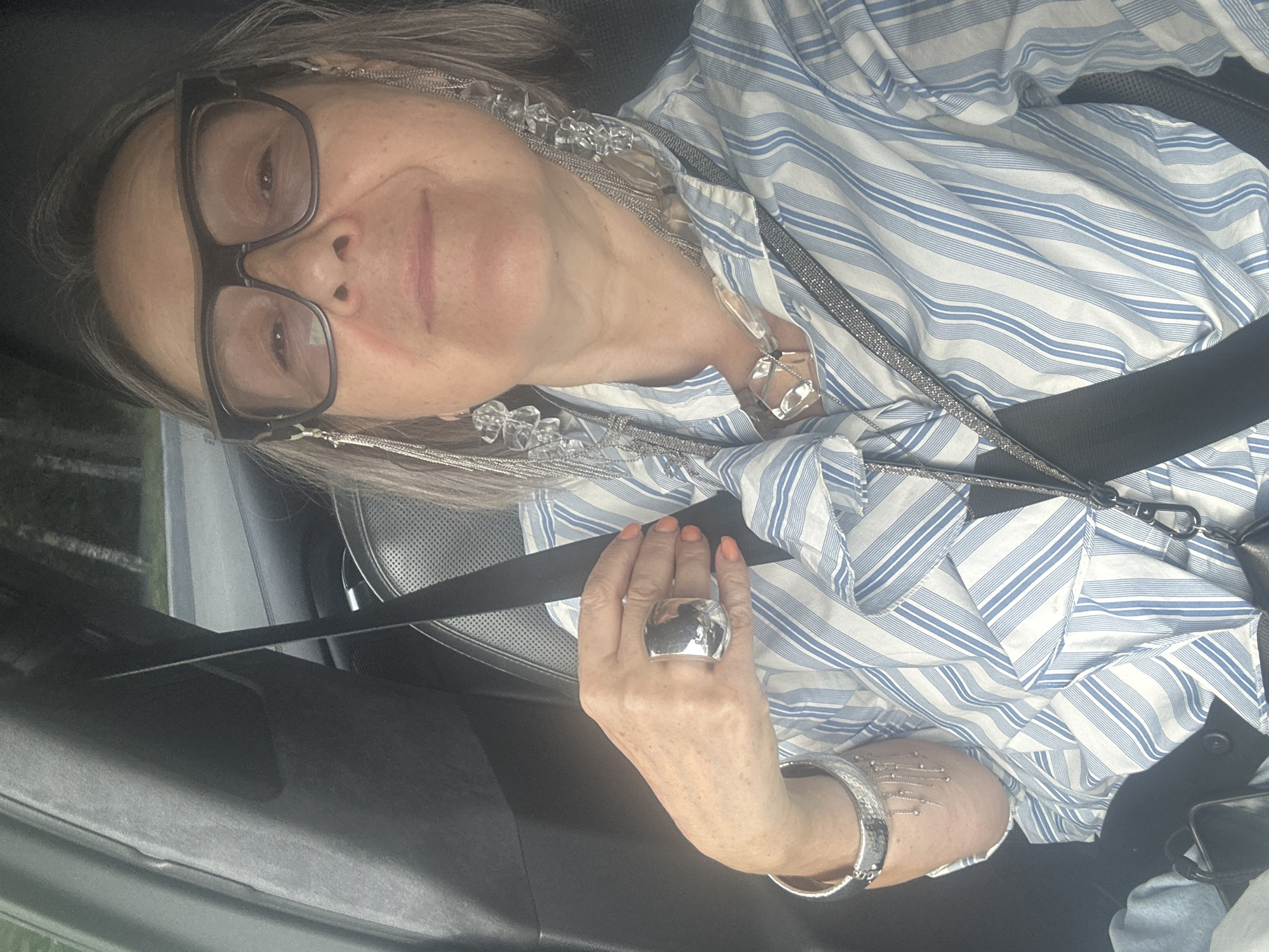 A woman wearing glasses and a blue and white striped shirt is in the driver's seat of a car. She is wearing a silver ring on her left hand and several silver necklaces and earrings. The seatbelt is across her chest. The background shows trees and a road. The time of day appears to be daytime.