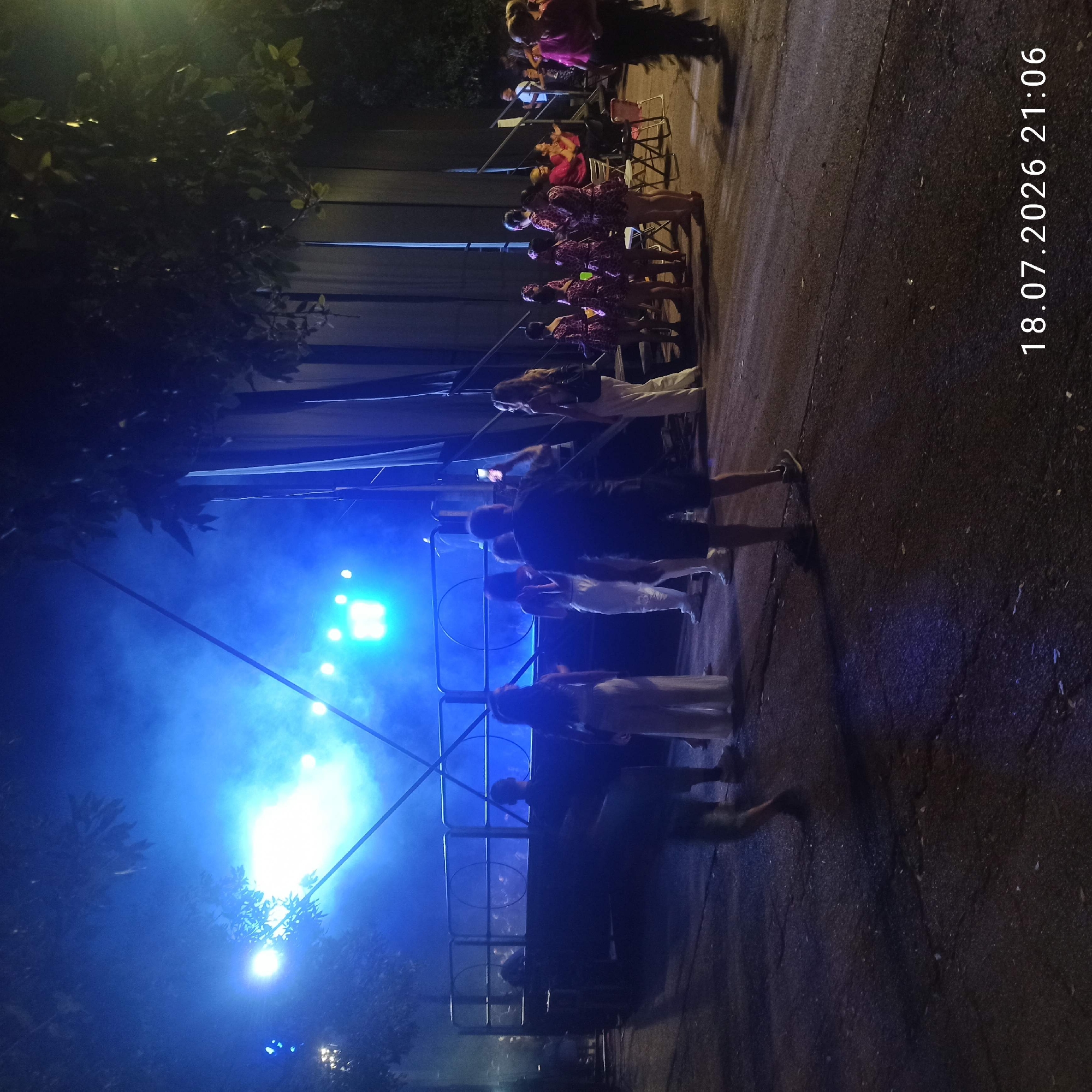The image depicts an outdoor nighttime event, possibly a performance or festival, taking place in the city of Bitonto, Italy. The scene is dominated by vibrant blue stage lighting and smoke, creating a dramatic atmosphere. In the foreground, several individuals are visible, with their backs mostly turned to the camera, suggesting they are audience members or participants. One person in the center foreground is taking a photo with a mobile phone. Further back, a stage or raised platform is set up, illuminated by bright lights, with more people on and around it. The background features dark trees and what appears to be a large, dark structure, possibly a building or stage backdrop. The ground is paved, with cracks and scattered debris visible. A timestamp in the lower right corner reads "18.07.2026 21:06".