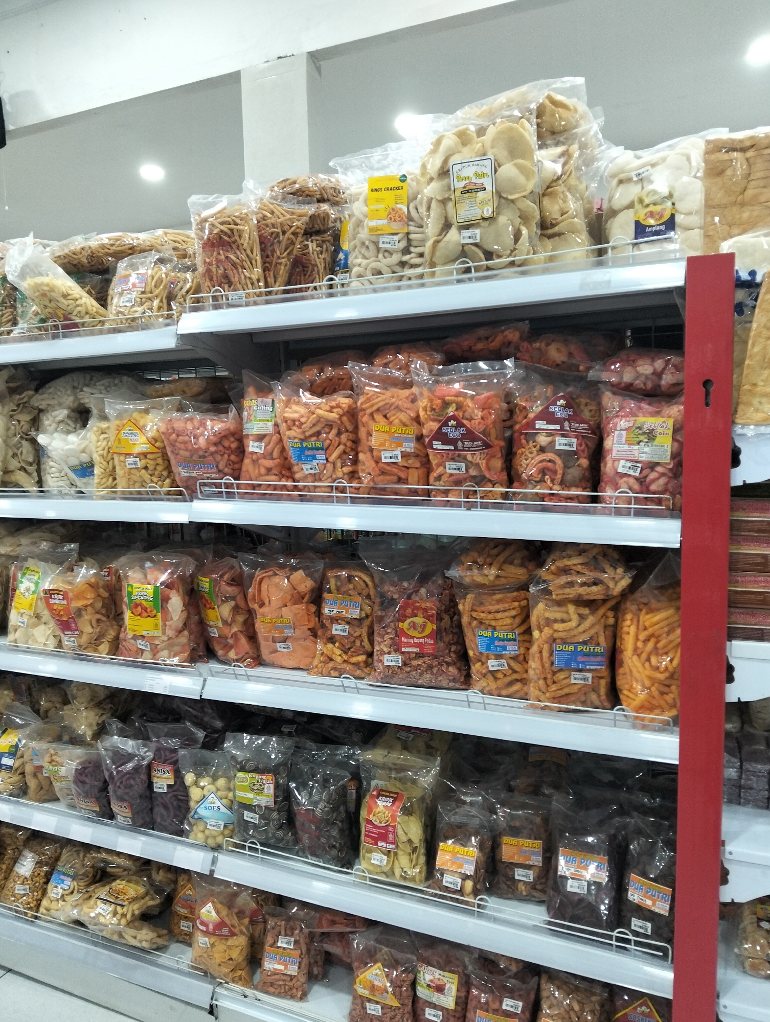 The image shows multiple shelves in a store filled with bags of snacks. The shelves are made of metal, and the snacks are packaged in clear plastic bags. The bags contain various types of snacks, including crackers, chips, and fried items, with different colors and shapes. Some bags have labels with text and images. A red vertical support beam is visible on the right side of the image. The overall environment appears to be a well-stocked snack aisle in a retail establishment, likely a minimarket or grocery store. The location is identified as Kedungrejo, Indonesia. There are no people or specific actions depicted in the image.
