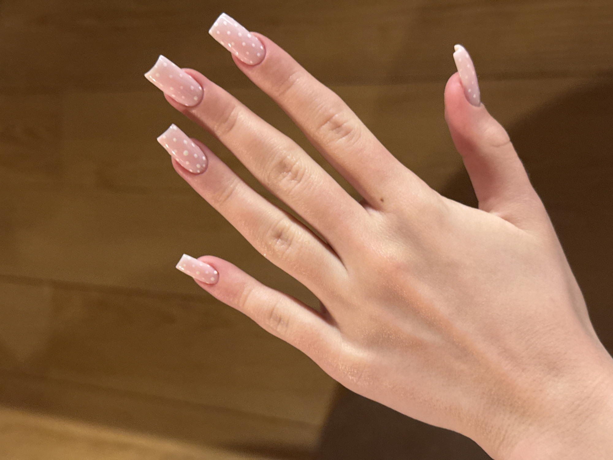 A hand with pink and white polka-dotted square-shaped nails is positioned against a wood surface. The hand is fair-skinned. No specific details indicate a location in Katun Bačko brdo, Montenegro.