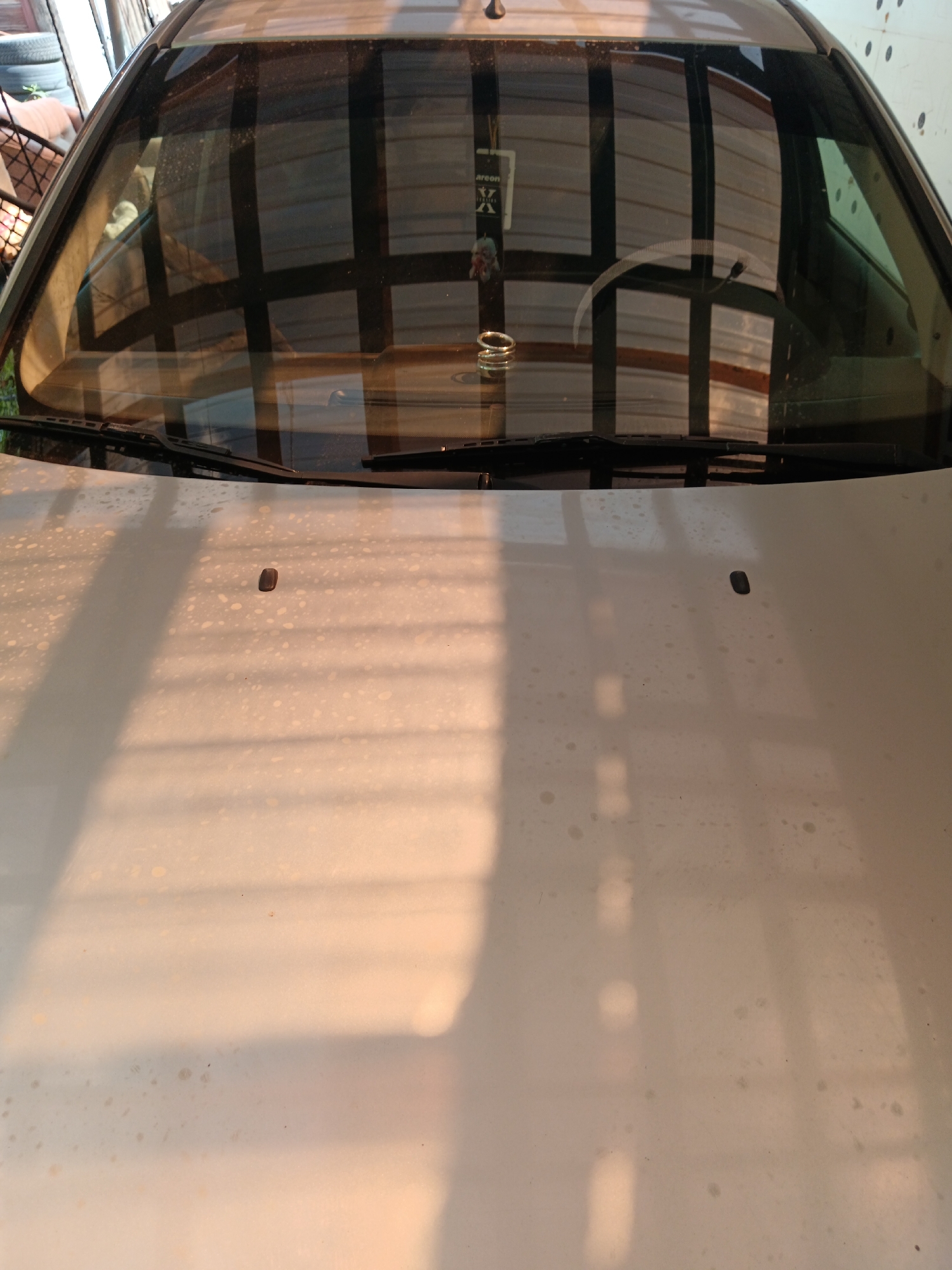 The image shows the hood and windshield of a silver car, likely parked outdoors in an urban or semi-urban environment. The car's surface is dusty and speckled with what appear to be water spots or light dirt. Sunlight casts strong, striped shadows across the hood, suggesting a covered area with openings, possibly a carport or a garage with a slatted roof. The reflections in the windshield are complex, showing a grid-like structure and possibly the interior of a building or a covered structure. A car air freshener hangs from the rearview mirror inside the car, and a coiled ring is visible on the dashboard. Two black windshield wiper blades are positioned on the lower edge of the windshield. The scene appears to be located in Sorochinsk, Russia, given the provided context.