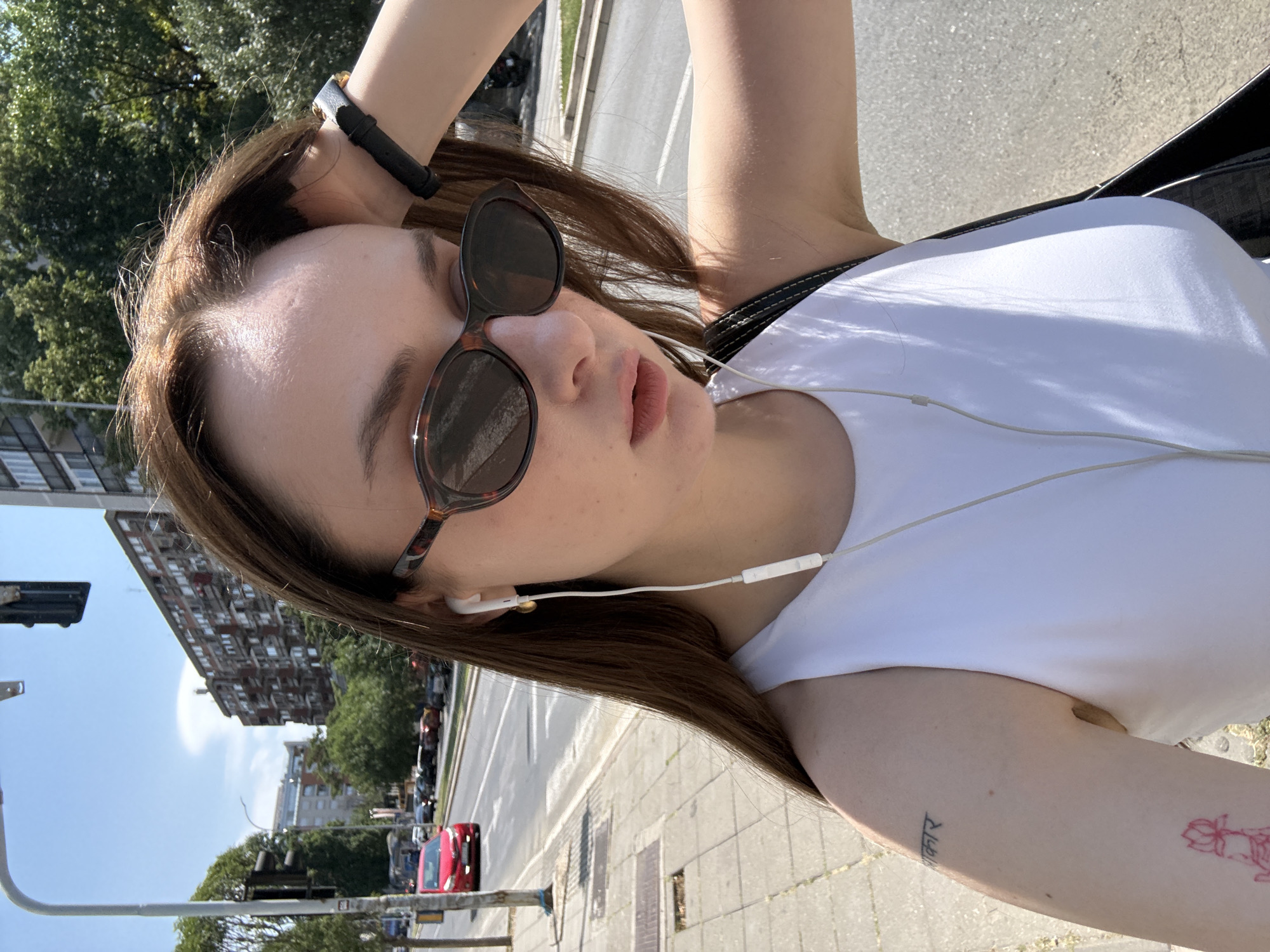 A young woman with long brown hair, wearing a white tank top, sunglasses, and white earbuds, takes a selfie outdoors on a sunny day. She is standing on a sidewalk next to a street in Belgrade, Serbia. Cars are visible on the road, along with buildings and trees in the background. The woman has a tattoo of what appears to be a lotus flower on her left arm and another tattoo above it with text in an Indian script. She is holding her right hand to her head.