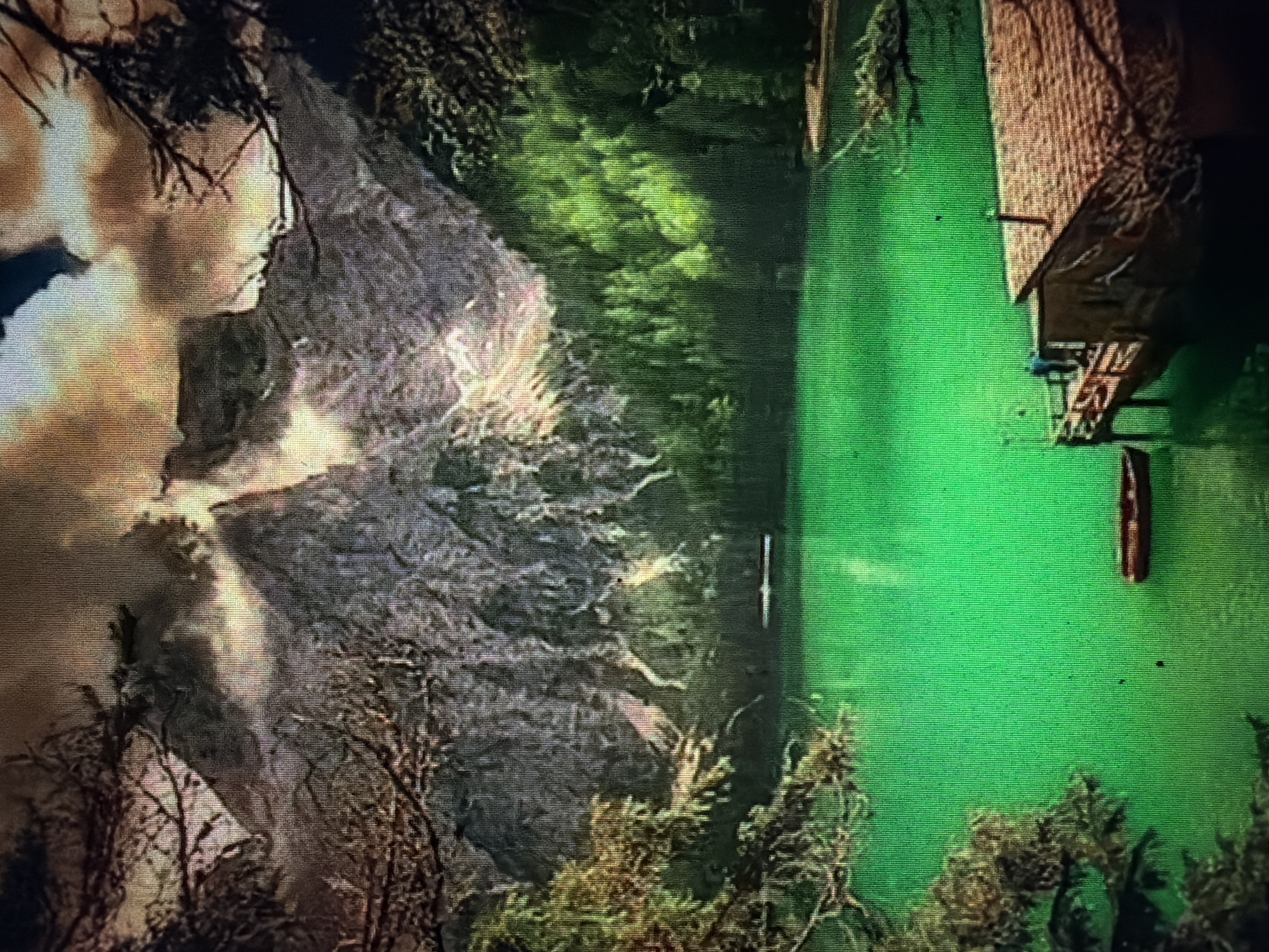 The image depicts a serene mountain lake scene. A vibrant green body of water occupies the foreground and midground, reflecting the surrounding scenery. In the background, towering, rugged mountains are partially obscured by dramatic, cloudy skies. Lush green pine trees line the shore of the lake, creating a dense forest. A wooden pier juts out into the water, with a small, rustic building constructed on it. Two figures, one wearing blue and the other appearing to be in silhouette, stand on the pier. A small, red boat is moored near the pier. The overall atmosphere is peaceful and natural, with the bright green of the lake and the rich greens of the forest contrasting with the grey of the mountains and the warm tones of the clouds. There is no discernible location within the city of Baan Mae Nam, Thailand depicted in this image.