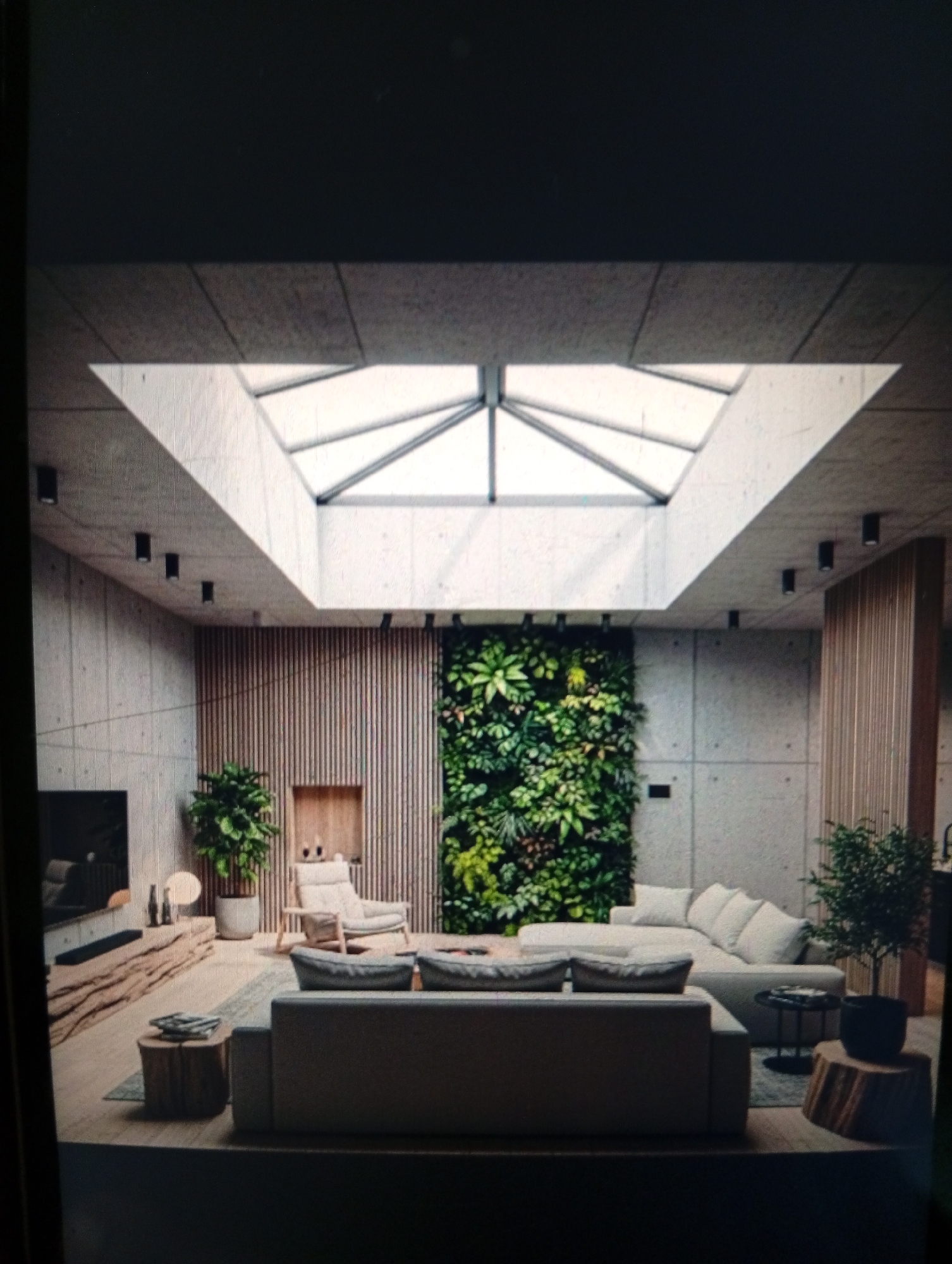 The image depicts a modern living room interior, seemingly in an upscale residential space. The room is characterized by a minimalist design with exposed concrete walls and a high ceiling featuring a large skylight. Dominating the center of the room is a large sectional sofa with light-colored cushions. A green wall consisting of various plants is a notable feature against one of the walls, providing a natural element. Other furniture includes a wooden console under a television, a lounge chair, and small side tables made of natural wood. The flooring appears to be a light-colored wood or tile. Overall, the aesthetic is clean, modern, and emphasizes natural light and greenery. There is no clear indication of the location being in Dutsen Alhaji, Nigeria, or any other city. There are no people shown in the image.