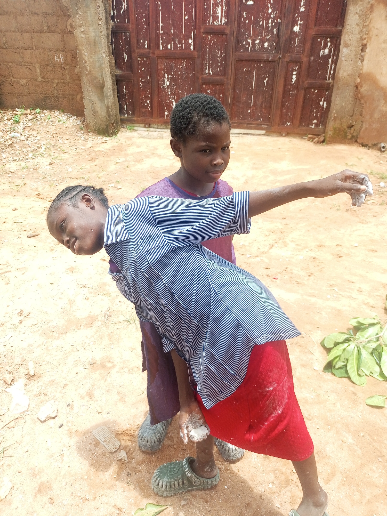 In Tempure, Nigeria, two young girls stand in a dusty, outdoor area. The girl in the foreground is bending backward, her body angled away from the viewer. She is wearing a blue and white striped shirt over a red skirt and dark green slip-on shoes. Her right arm is extended, and her hand is covered in a white substance, possibly chalk or mud. The girl behind her, looking towards the camera, is wearing a purple shirt and also dark green slip-on shoes. Behind them, a large, weathered, dark red double door stands out against a brick wall and a sandy, uneven ground with scattered debris and some green foliage. The overall scene suggests a casual, perhaps playful, moment in a rural or semi-urban setting.