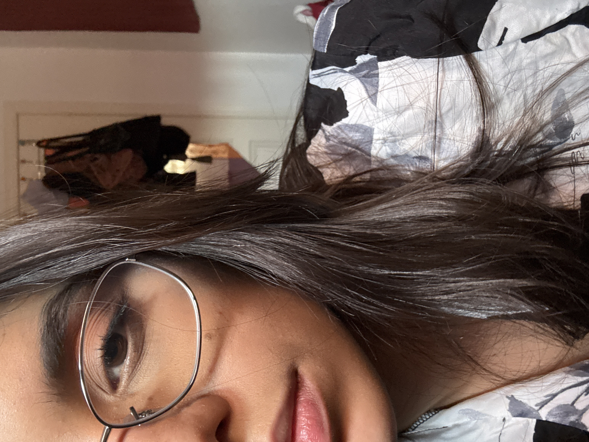 The image is a close-up portrait of a woman's face, cropped to show her right eye, nose, lips, and part of her hair and clothing. She is wearing glasses with thin, silver frames. Her hair is dark brown and falls loosely around her face. She is wearing a black and white patterned shirt. The background is out of focus, but appears to be an indoor setting, possibly a bedroom, with items hanging on a wall and a door visible. The lighting suggests it is daytime. The image does not contain any information that would allow for determination of its location in Las Rozas de Madrid, Spain.