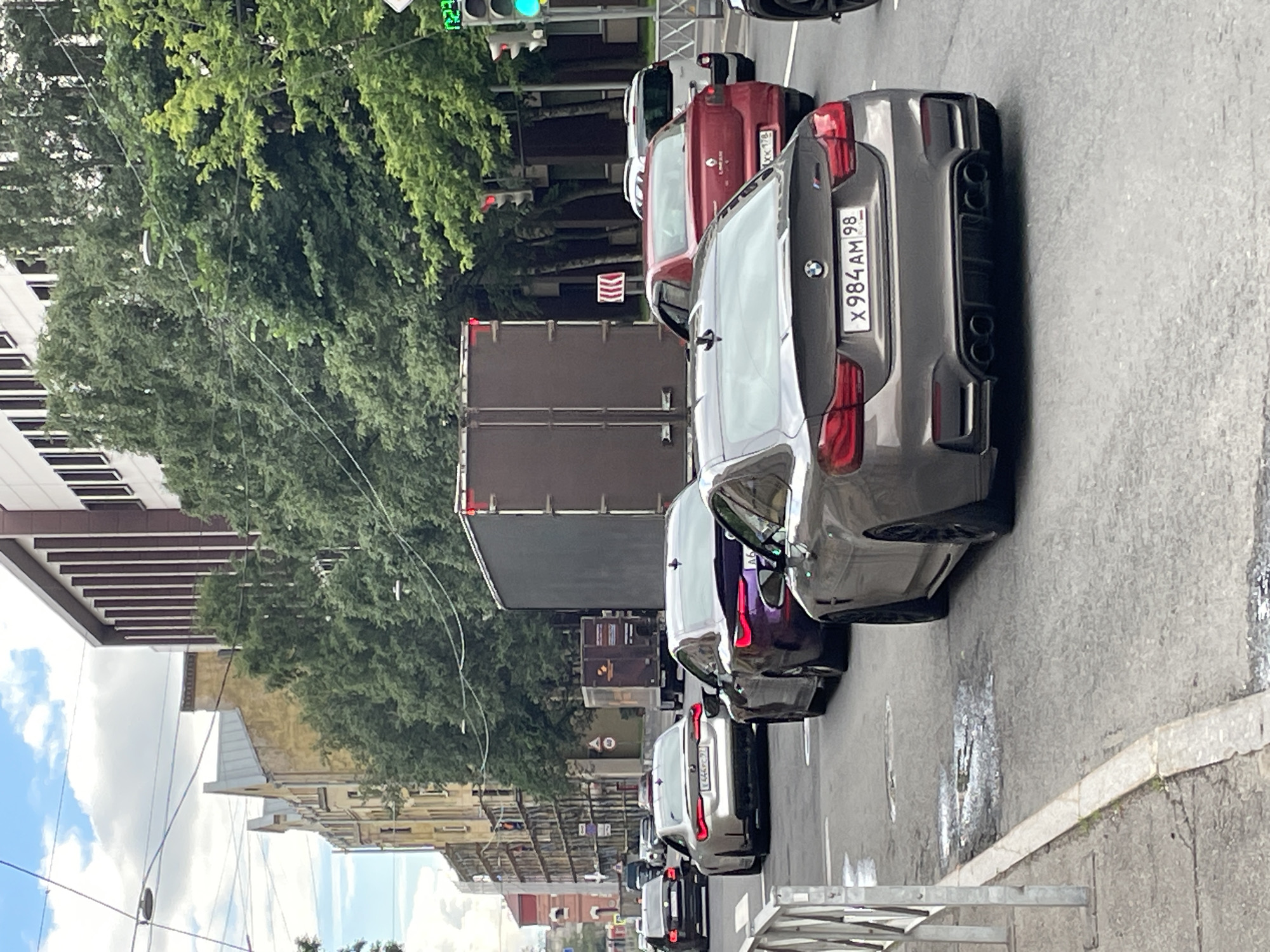 This is a street-level, medium-angle, slightly over-the-shoulder shot taken on a cloudy day in Saint Petersburg, Russia, depicting a traffic jam on a multi-lane road. The foreground is dominated by the rear of a dark grey BMW M4 coupe with a Russian license plate X984AM98. To its left, a portion of a light silver BMW coupe is visible. Further in the background, a dark purple sedan is also in traffic, followed by a red sedan and a white SUV. A large, dark brown truck with a trailer is positioned to the left of the purple sedan. In the background, there are several multi-story buildings with classical and modern architectural styles, some with visible construction or scaffolding. Trees line the street, and power lines are strung across the sky. Traffic lights are visible, with one showing green. The road surface appears wet in places.