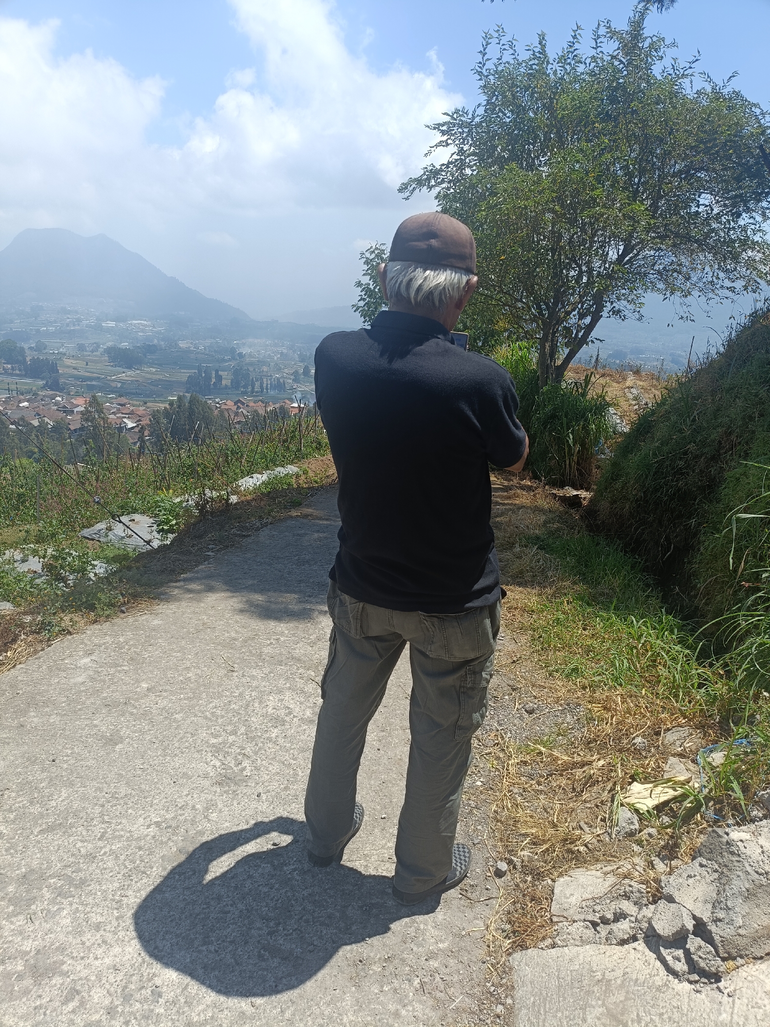 A person stands with their back to the camera, looking out over a valley. They are wearing a dark-colored polo shirt, olive-colored cargo pants, a brown baseball cap, and checkered shoes. The person's arms are crossed, and they are holding a smartphone in their left hand. The scene appears to be a scenic overlook in or near Kopeng, Indonesia, with a lush green tree to the right of the person. In the background, a mountain range is visible, shrouded in mist, with a settlement nestled in the valley below. The ground is a paved path with dry grass and some rocks. The sky is blue with scattered clouds. The lighting suggests it is daytime.