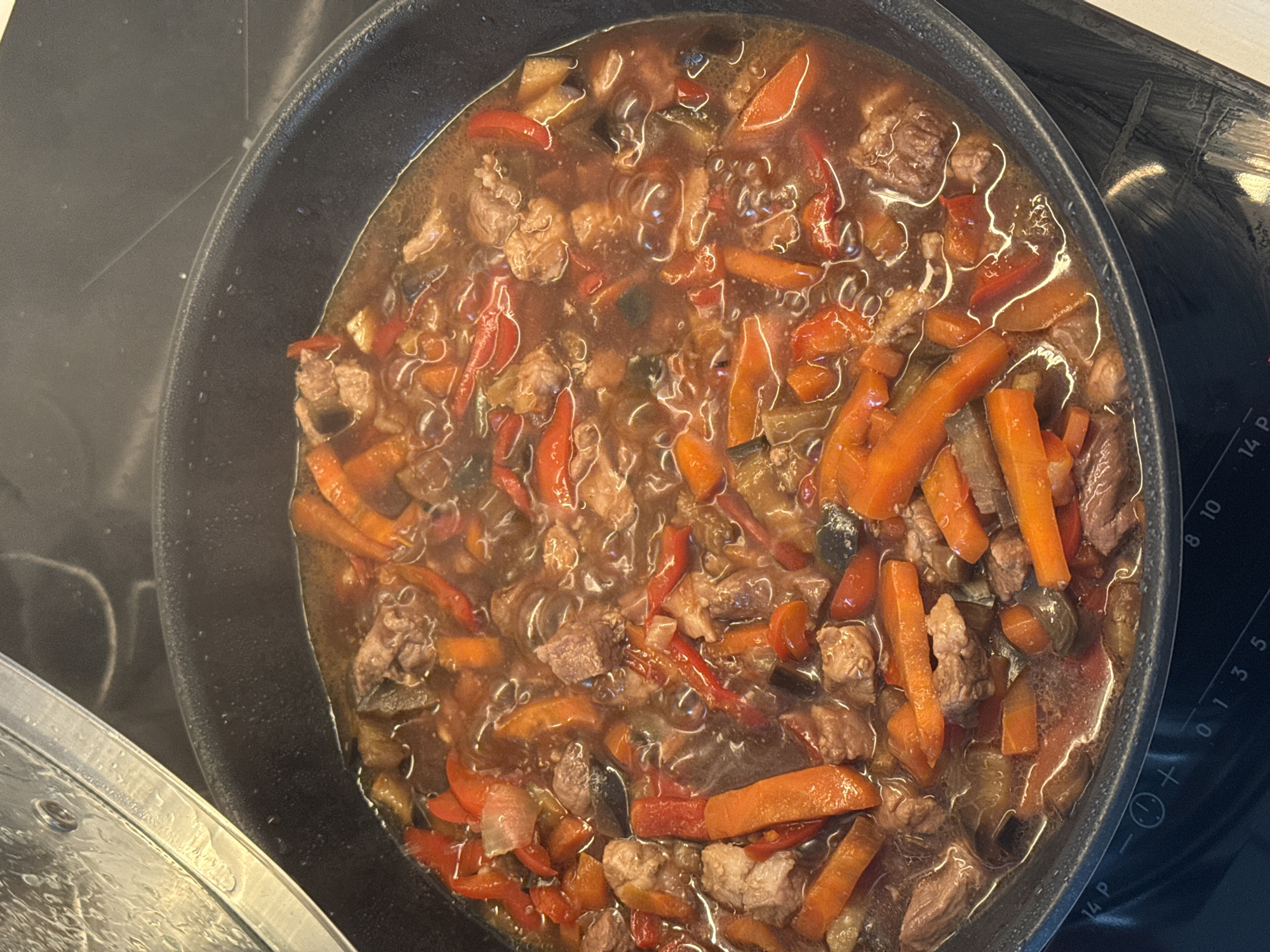 An overhead, close-up view captures a hearty stew simmering in a dark, round pan on a black induction or ceramic stovetop in a kitchen in Shirkovo, Russia. The pan is filled with a rich, dark brown liquid, gently bubbling, suggesting it is actively cooking. Faint steam is visible rising from the dish.
The stew contains numerous ingredients: prominent chunks of tender meat, likely beef or pork; vibrant orange carrot sticks cut into elongated pieces; and bright red bell pepper strips. Darker, softer vegetable pieces, possibly eggplant or zucchini, are also mixed throughout the bubbling liquid.
To the bottom left, a clear glass pot lid, covered with condensation, is partially visible, resting next to the pan. On the right side of the stovetop, a control panel with white markings is visible. These markings include numbers "0", "1", "3", "5", "8", "10", and "14 P", which likely denote heat levels or timer settings. A circular icon featuring a plus and minus sign, and another small circular icon, are also present on the control panel. The scene conveys the process of preparing a home-cooked meal.