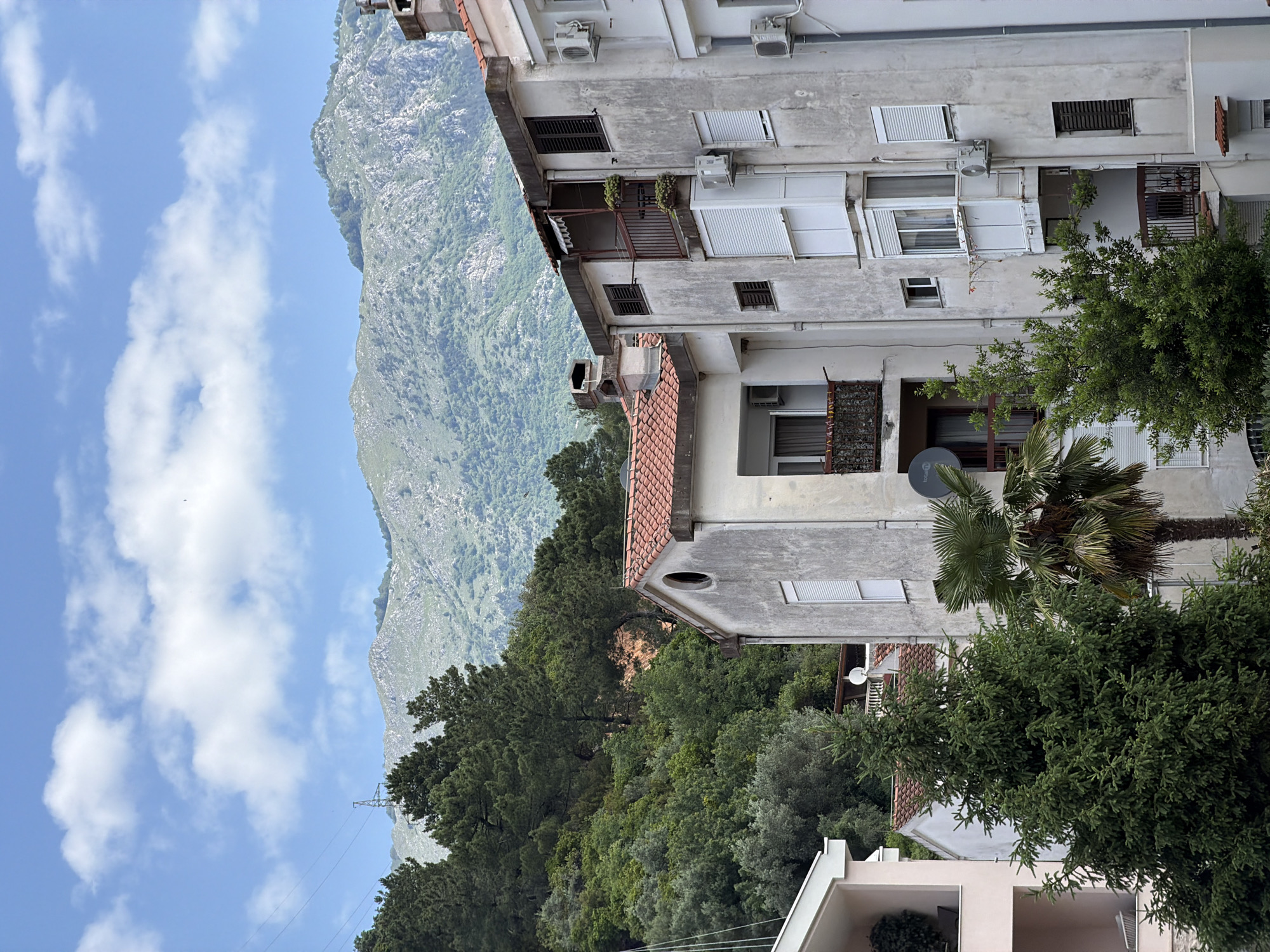 The image depicts a partially obstructed view of a mountain landscape in Budva, Montenegro, juxtaposed against residential buildings. The sky is visible with scattered clouds. The mountain is prominent, with a rugged terrain covered in sparse vegetation. To the right of the mountain, a weathered, multi-story building dominates the frame. The building features a mix of closed and open windows, some with shutters. Several air conditioning units are attached to the exterior walls. A variety of trees and greenery are visible between the buildings. Utility lines are visible crossing the sky, adding to the urban-nature contrast. A satellite dish is visible on the building.