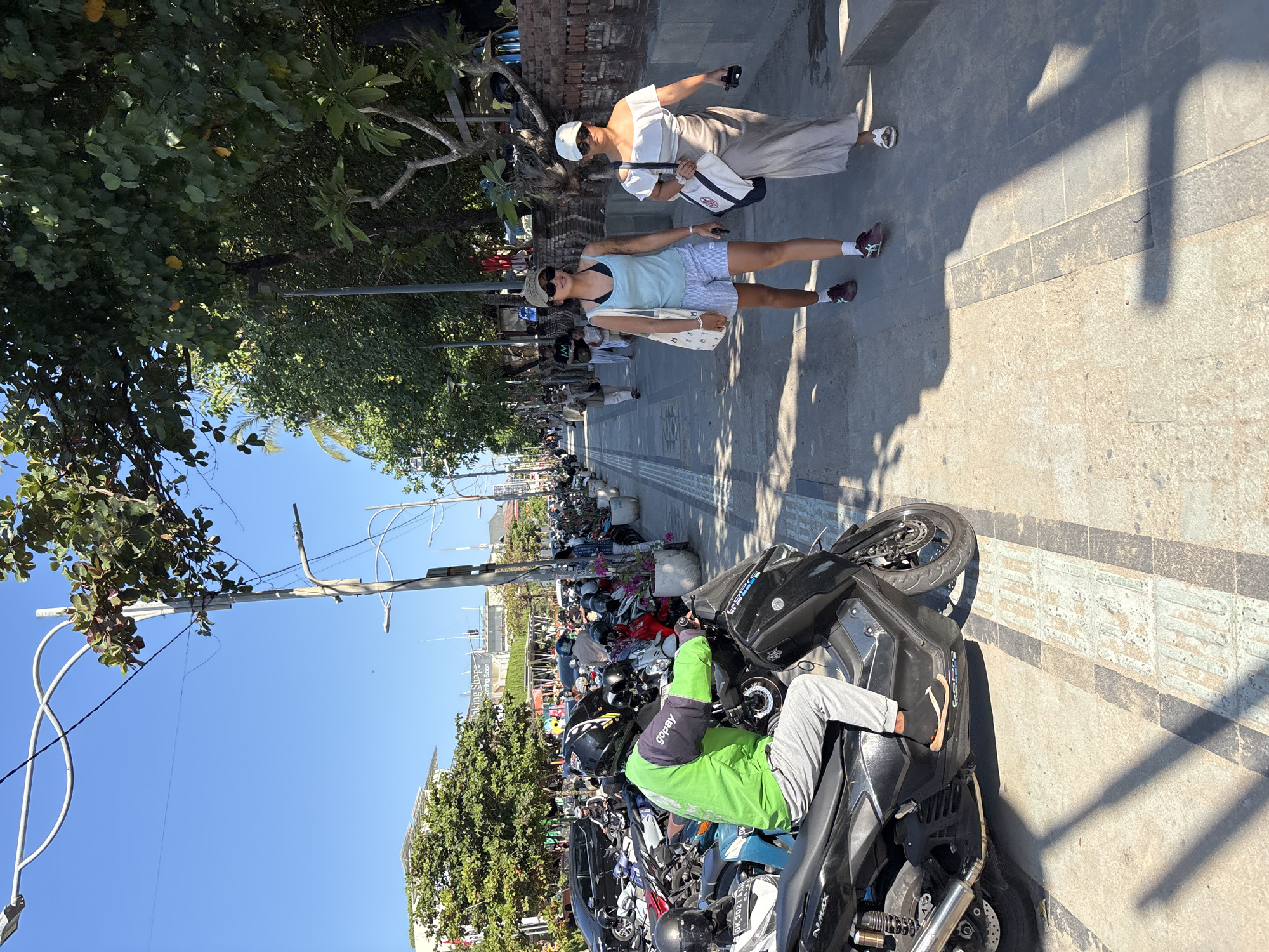 In Kuta, Indonesia, a street scene unfolds under a bright, clear sky. A person wearing a green jacket with "Gopay" branding rides a black scooter, parked alongside a row of other motorcycles. The pavement is a textured mix of grey and beige tiles, with a tactile paving strip running along its edge. To the right, two women walk side-by-side on the sidewalk, carrying tote bags. The woman on the left wears a light blue tank top and striped shorts, while the woman on the right is dressed in a beige off-the-shoulder dress. Beyond the sidewalk, a brick wall and lush green trees line the path. In the background, buildings and more trees suggest a bustling urban environment.