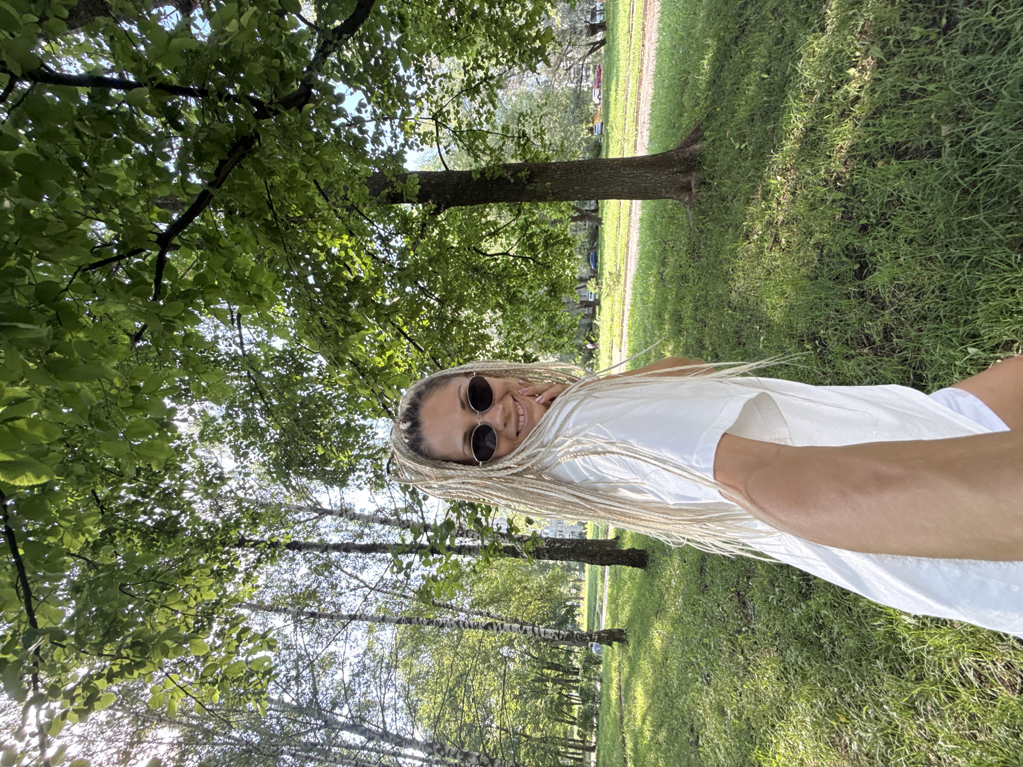 A woman with blonde dreadlocks and sunglasses is taking a selfie in a park. She is wearing a white t-shirt and white shorts. The park has green grass, trees, and a paved walkway in the background. The woman is smiling and her right hand is touching her chin. The location is Rusanovo, Russia.
