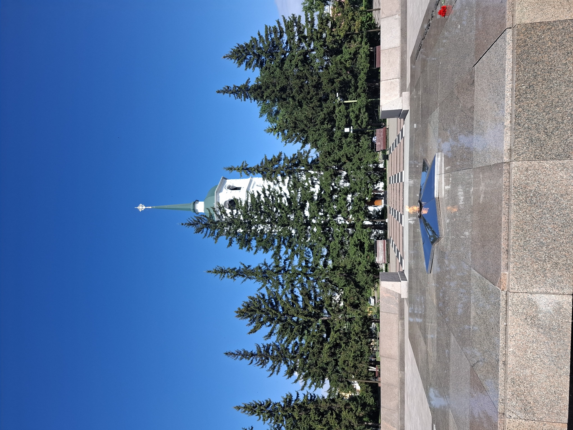 The image depicts a view of the Eternal Flame memorial in Irkutsk, Russia, on a clear, sunny day with a bright blue sky. In the foreground, a large, polished granite structure surrounds a star-shaped eternal flame, which is burning and reflecting in the polished surface. Behind the memorial, a line of tall evergreen trees stands in front of a white building with a green spire and a cross at its peak, likely a church or a bell tower. Benches are visible in the background, and a few people are seated on them. The overall scene conveys a sense of solemnity and remembrance.