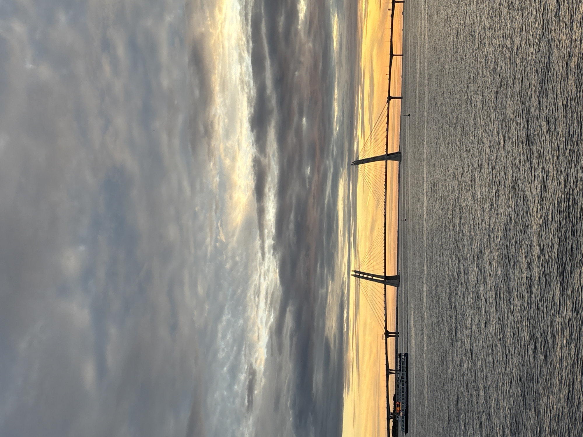 The image depicts a view of the Western High-Speed Diameter (WHSD) bridge in Saint Petersburg, Russia, during a sunset. The sky is filled with dramatic clouds, ranging from dark gray to lighter shades of gray and yellow, illuminated by the setting sun. The water in the foreground is calm with gentle ripples.
A large cable-stayed bridge with distinctive towers is the central focus, spanning across the water. To the left of the main bridge, another section of the WHSD bridge is visible, supported by pylons. A passenger ship, identified by its white hull and multiple decks, is sailing on the water towards the left of the frame, passing under the bridge structure. Several small buoys are scattered on the water. The scene captures a moment of twilight, with the bridge illuminated and reflecting on the water.