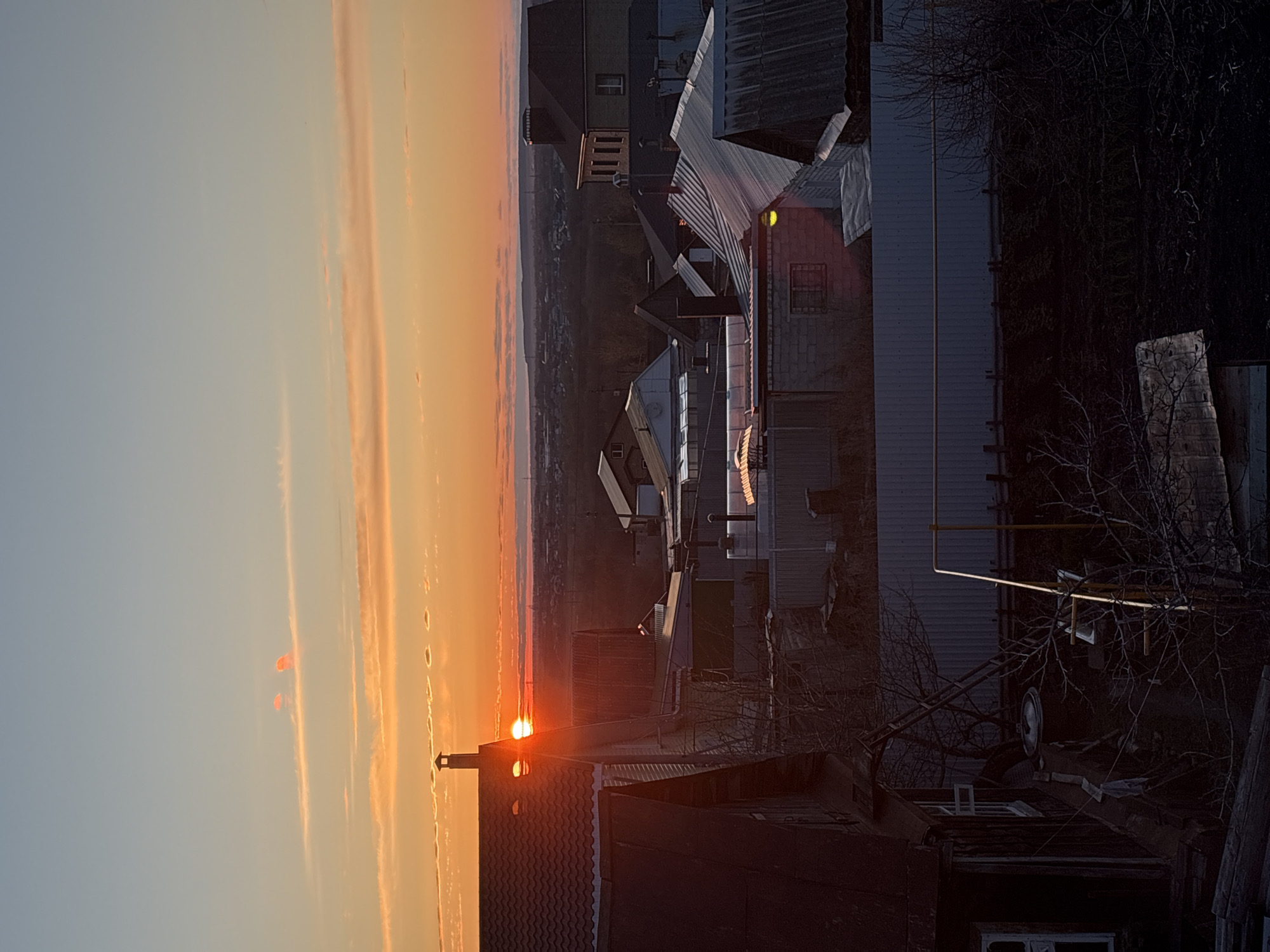 The image displays a vibrant sunset over an urban landscape, likely captured from an elevated vantage point in Ulyanovsk, Russia. The sky, occupying the left side of the vertically oriented image, transitions from a pale blue at the top to a brilliant orange and yellow hue towards the right, where the sun is positioned. The sun, a luminous, partially obscured orb, is low on the horizon, casting a strong golden light. Several horizontal streaks of illuminated clouds traverse the sky, reflecting the intense colors of dusk.
The right side of the image is dominated by a dense cluster of buildings, mostly appearing in deep silhouette or shadow due to the backlighting from the setting sun. In the immediate foreground, a dark tiled roof with a prominent chimney stands out, directly beneath the sun. Further into the midground, various buildings with corrugated metal and other roof types are visible, their forms indistinct in the low light. Some structural details, such as windows, are discernible, and one building emits a soft yellow glow from an internal light source.
Bare, leafless trees and dark, dense vegetation are scattered throughout the scene, particularly on the far right, suggesting a colder season like late autumn or winter. Details within the shadowed areas include pipes running along a building's exterior and what appears to be a ladder and a car wheel nestled within the dense foliage. The distant horizon reveals a faint, hazy landscape. The overall mood is quiet and atmospheric, with no human activity or visible text.