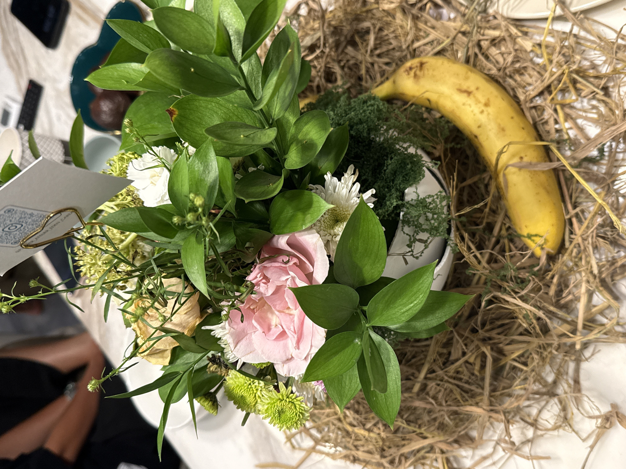 The scene presents an indoor arrangement featuring a bouquet of flowers with lush green foliage, interspersed with pink, white, and green blooms, nestled in a white container. To the right of the arrangement, a banana lies on a bed of dry hay-like material. The overall setting is a table surface, with glimpses of other objects such as tableware and what appears to be part of a menu card with a QR code. The location is presumed to be Kerobokan, Indonesia.