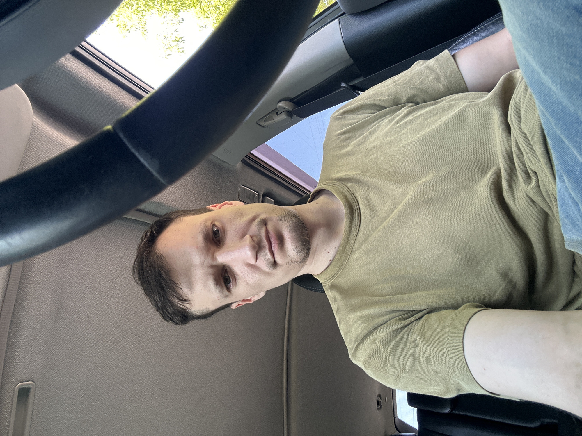 A selfie taken from the perspective of someone sitting in the driver's seat of a car. The person, a man with short brown hair and a slight stubble, is looking directly at the camera with a neutral expression. He is wearing a plain olive-green t-shirt and blue jeans. The steering wheel of the car is visible in the foreground, partially obscuring the view. The car's interior, including the ceiling and a portion of the seat, is visible. Through the window, trees and a bright sky suggest an outdoor, daytime setting. The specific location in Makiv, Ukraine, is not discernible from the visual information provided.