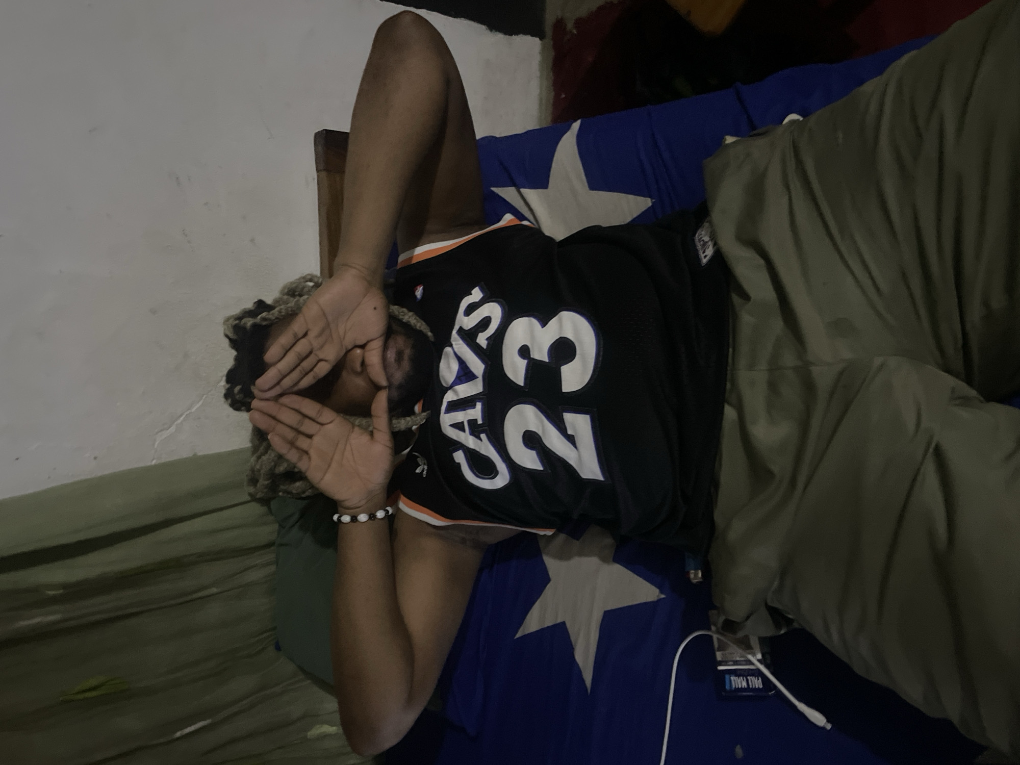 A person lies on their back on a bed. The person is wearing a "Cavs 23" basketball jersey and khaki pants. Their face is covered by their hands. They have beaded braids. The bed has blue sheets with white stars. A pack of Pall Mall cigarettes and a phone charger are next to the person on the bed. The background appears to be a room with painted walls. There is no indication of the specific location of Jos, Nigeria.