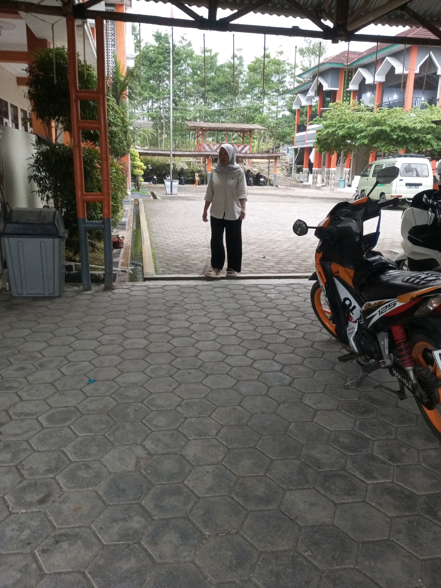 In Kalibeber, Indonesia, a woman wearing a light-colored hijab and a long-sleeved shirt with dark pants stands on a paved walkway. She is facing away from the camera, looking towards a building. A motorcycle, predominantly orange and black with "Honda" and "125" visible on its side, is parked in the foreground, angled towards the right. Behind the woman, a white van is parked near the building. The environment appears to be an outdoor area adjacent to a building with orange and white accents, surrounded by trees. The ground is covered in hexagonal paving stones. A grey trash can is positioned to the left of the walkway.