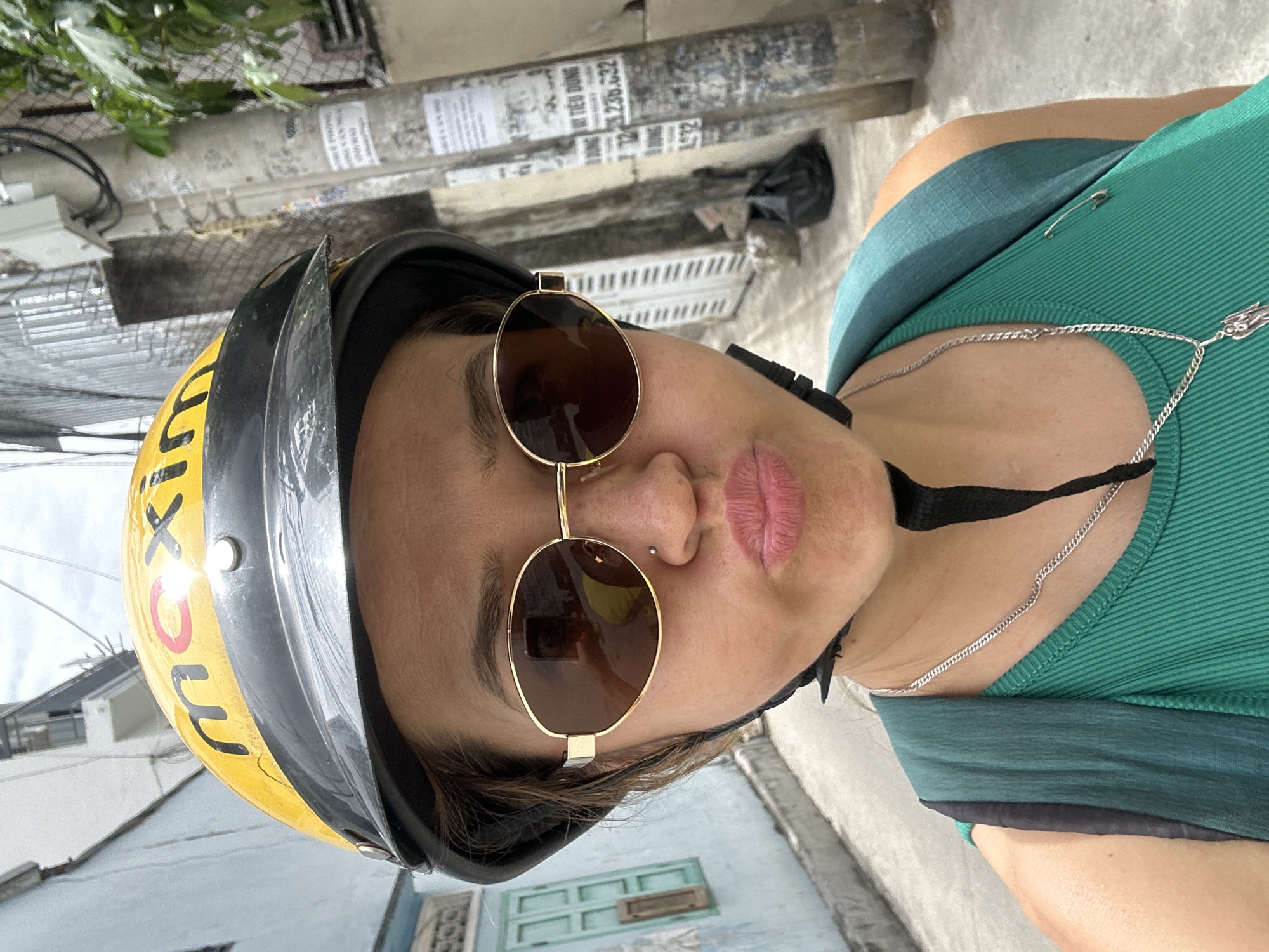 A person wearing a yellow motorcycle helmet with the word "maximo" written on it and gold rimmed sunglasses is oriented in the center of the image. The person is also wearing a green shirt. The background shows a residential street, including a light blue building with aqua-colored doors, as well as concrete utility poles with attached signs. The setting is most likely in a city, possibly the Bac Nha Trang Ward in Vietnam.