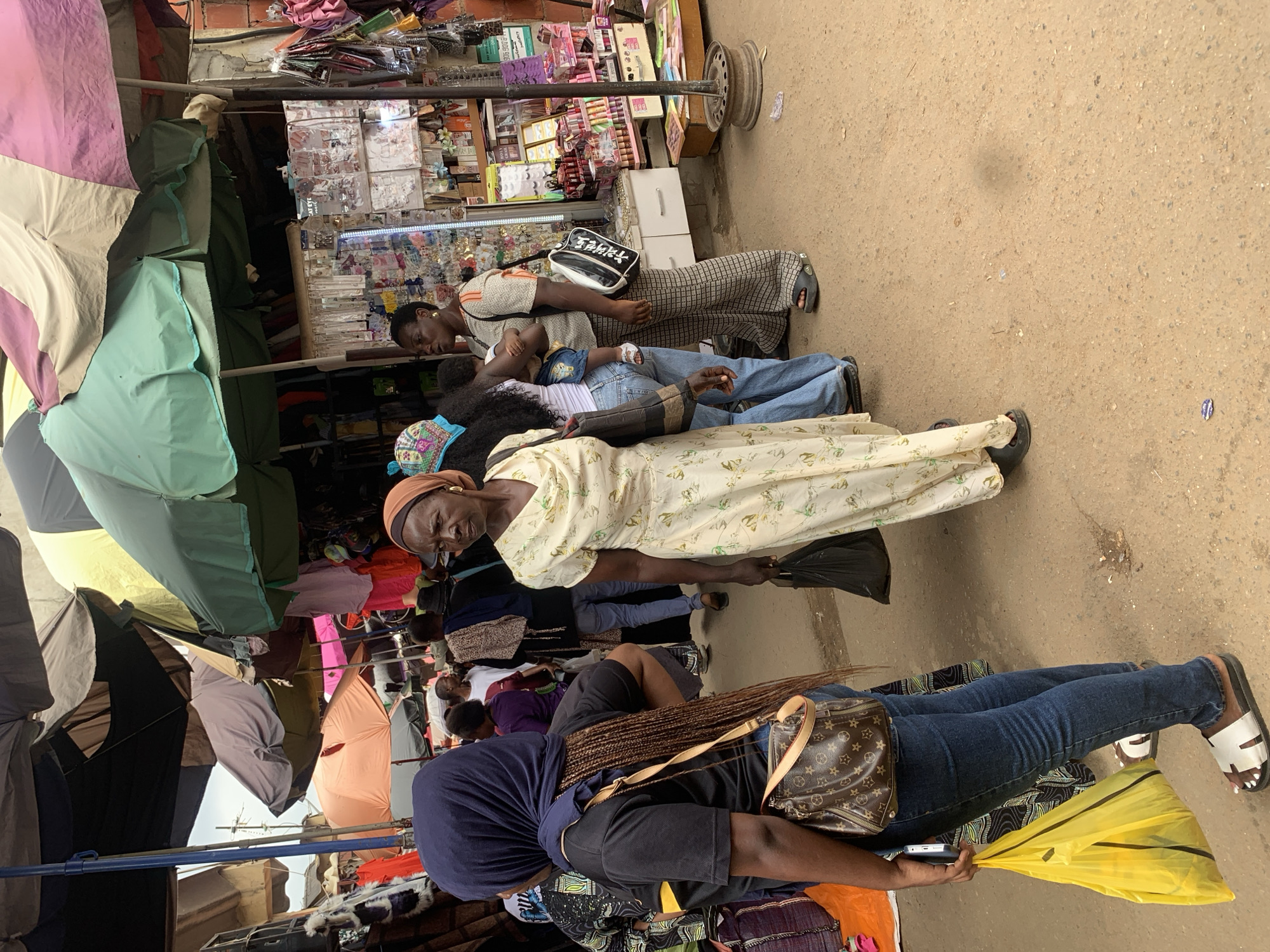 A group of people are gathered at an outdoor market in Abuja, Nigeria. The scene is bustling with activity, suggesting it is a busy market day. Several individuals are visible, including women in traditional clothing and one person carrying a child. The setting appears to be a street market with various stalls set up, displaying goods like fabrics, umbrellas, and possibly cosmetics or accessories. Colorful umbrellas are prominent, providing shade over the stalls and the shoppers. The ground is dusty, indicating a dry environment. The time of day is unclear, but the bright light suggests it could be daytime, possibly late morning or afternoon. The overall mood seems to be one of everyday commerce and community interaction. Some of the stalls display neatly arranged products, hinting at an organized market setup. The presence of a young child being carried adds a domestic element to the scene. No specific text is clearly legible in the image, although there might be some labels on the products displayed at the stalls.