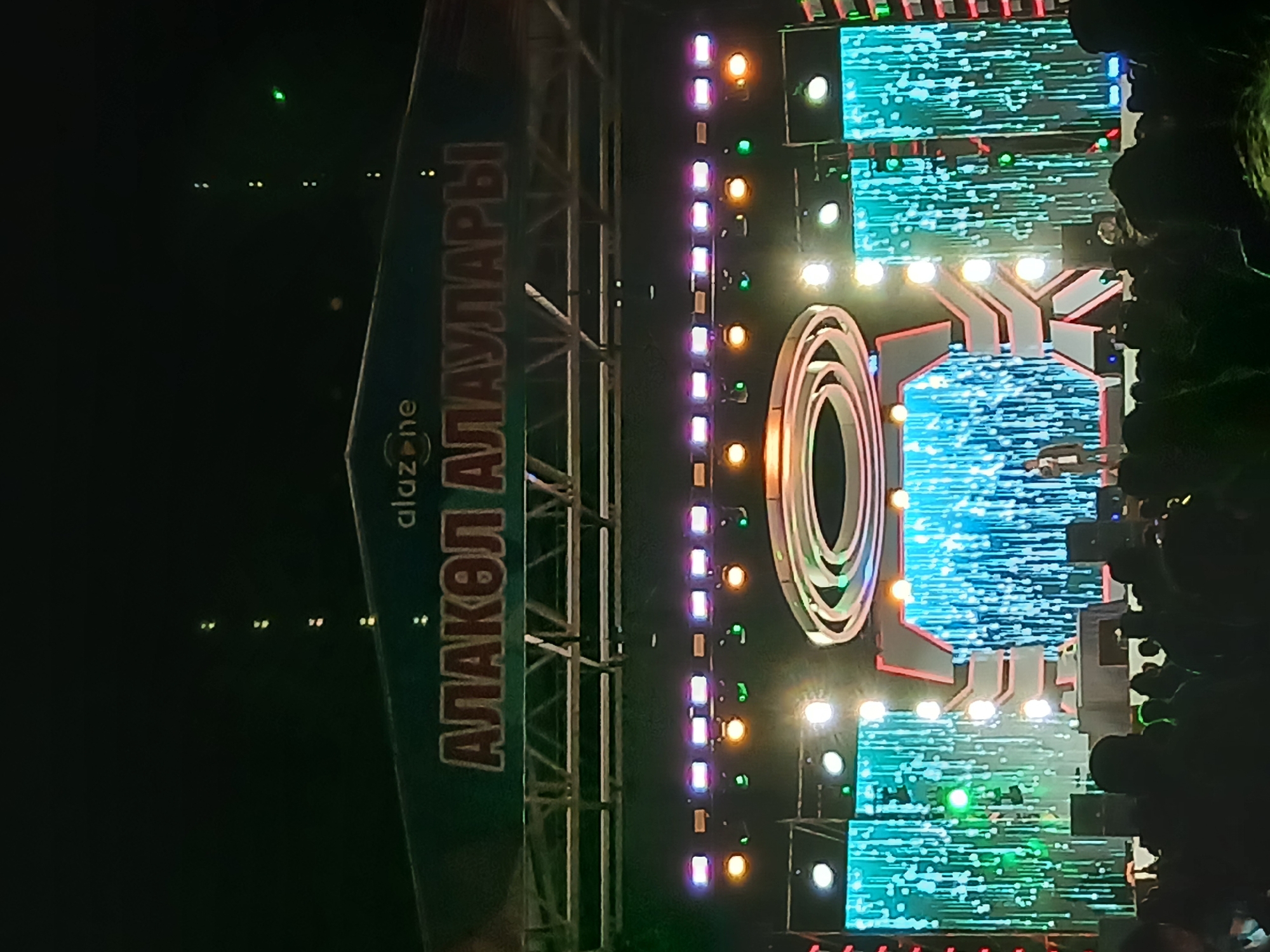 The image shows a night scene at an outdoor event, likely a concert or festival, with a large stage set up. The stage features prominent lighting, including rows of purple and pink stage lights, and numerous yellow and green spotlights. A large, illuminated sign with Cyrillic lettering, appearing to read "АЛАКӨЛ АЛАУЛАРЫ", is displayed above the stage, along with a smaller logo that says "alazone". The stage backdrop is a large LED screen displaying a vibrant, cascading blue and green light pattern, resembling falling rain or digital particles. In the foreground, a silhouetted crowd of people is visible, facing the stage. One individual, dressed in a dark jacket and trousers, stands on the stage in the spotlight. The overall atmosphere suggests a lively public gathering. The context of Zharbulak, Kazakhstan is not visually evident in the provided image.