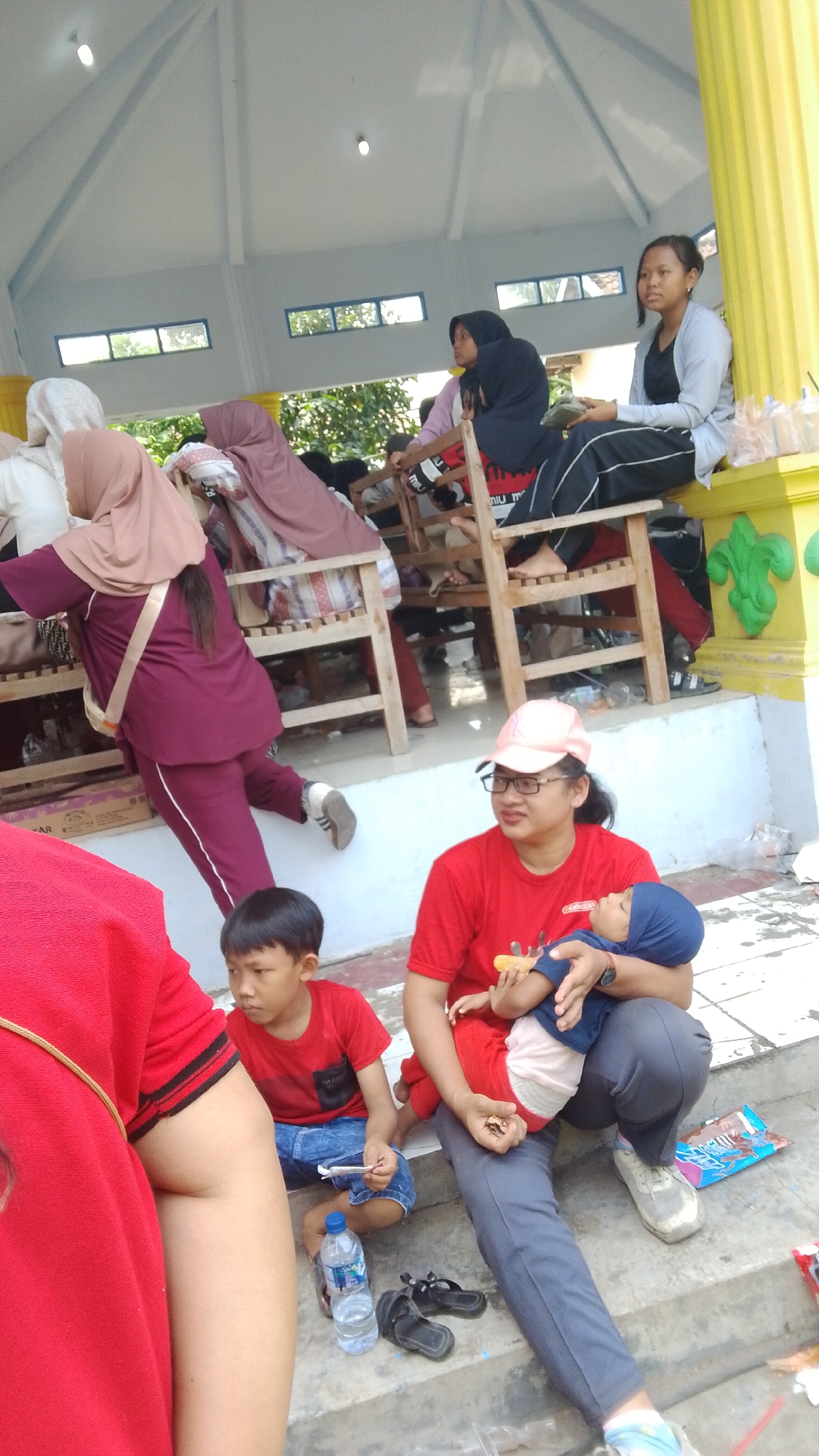 In Mancar, Indonesia, a group of people are gathered outdoors under a white, sloped roof structure. In the foreground, a woman in a red shirt and grey pants holds a baby wrapped in a blue cloth. To her left, a young boy in a red shirt and blue shorts sits on concrete steps, holding a white object. In the background, several other individuals, mostly women wearing hijabs, are seated on wooden benches. The environment appears to be a public space, possibly a community gathering area or a park, with some decorative elements on a yellow pillar.