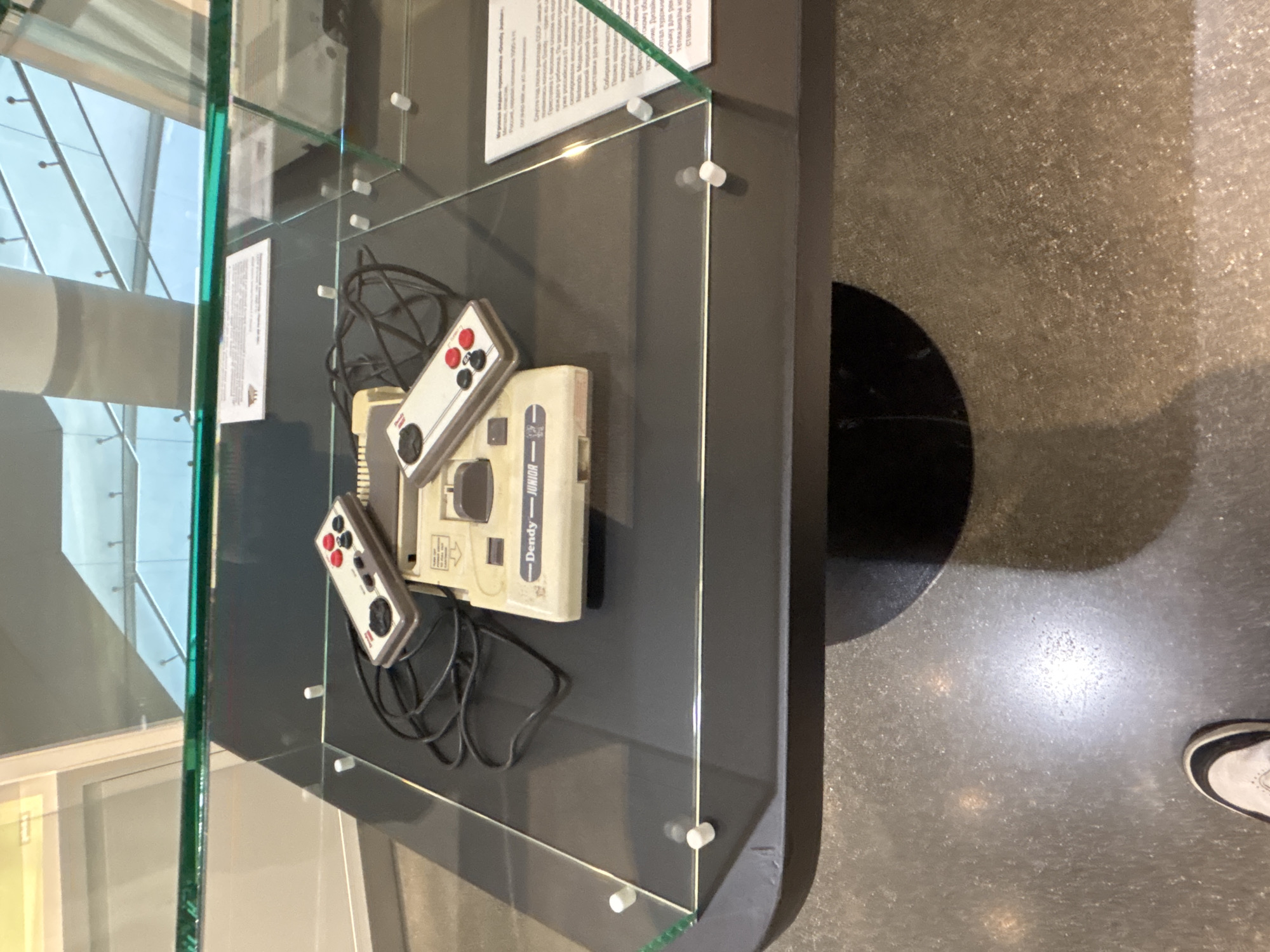 A Dendy Junior game console and two associated controllers are displayed within a clear glass exhibition case atop a dark grey, rounded rectangular plinth. The console, a beige unit with "Dendy — JUNIOR" printed on its front, is positioned centrally. Two identical beige controllers, featuring black D-pads and red and black action buttons, rest on either side of the console, connected by coiled black wires. White cylindrical supports secure the glass panels of the display case. Two white informational placards, displaying text primarily in Cyrillic, are visible beneath the glass on the right side, with one partially obscured behind the console. The reflections in the glass reveal a brightly lit, modern indoor environment with high ceilings and architectural elements. The floor, visible in the lower portion of the image, is a textured grey surface. A person's left foot, wearing a white and black shoe, is partially visible at the bottom edge of the frame, indicating an observer. This scene is located in a museum or exhibition hall in Yekaterinburg, Russia, likely the Yeltsin Center Museum or a similar institution focused on technology or cultural history.