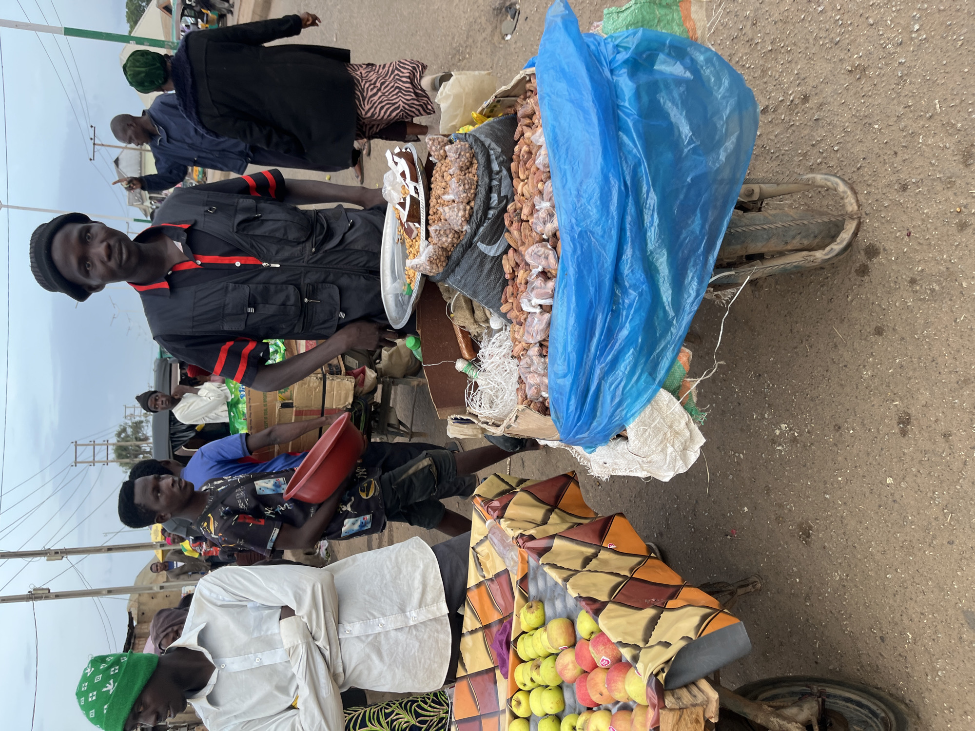 A street market scene is depicted in Bukuru, Nigeria, with several people and food stalls visible. In the foreground, a man wearing a black vest with red trim and a black beanie stands looking towards the viewer. To his left, another man in a white long-sleeved shirt and a green beanie stands with his arms crossed. In front of them, a cart displays a variety of fruits, including apples, arranged on a colorful patterned cloth. Beside this, another cart is filled with nuts and dried fruits, partially covered by a large blue plastic sheet. Other individuals can be seen in the background, some tending to stalls or walking through the market. The environment appears to be an open-air market with power lines and some structures in the distance. The ground is a dusty, reddish-brown dirt.