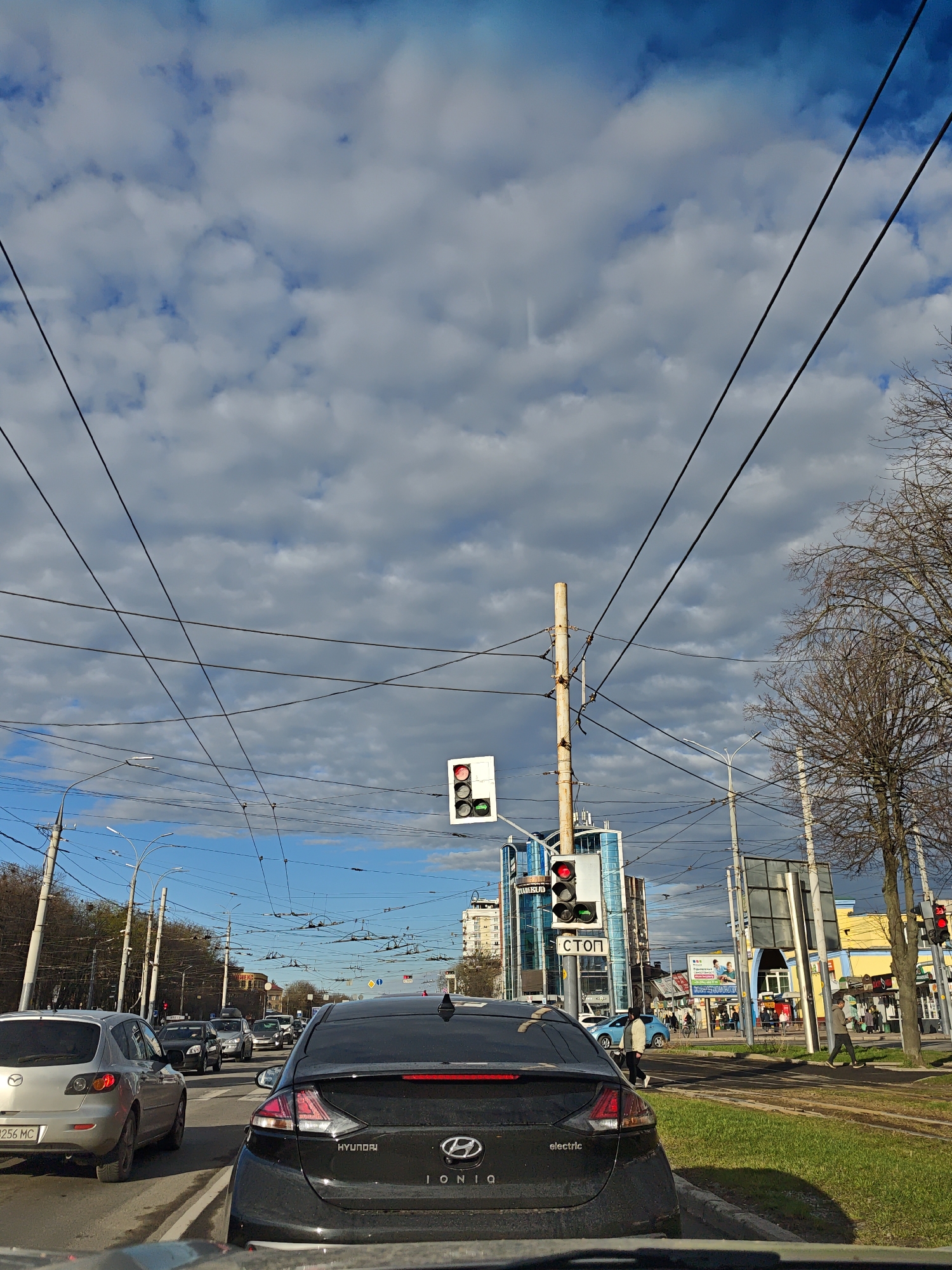 The image depicts a street scene in Vinnytsia, Ukraine, with a focus on traffic stopped at a traffic light. The sky is predominantly blue with scattered, puffy clouds. Overhead wires are visible crisscrossing the sky. In the foreground, the rear of a black Hyundai Ioniq electric car is prominent, followed by other cars in the traffic queue. The traffic light ahead shows a red light. Pedestrians are visible on the sidewalk to the right of the road. Buildings, including a modern glass structure and older, colorful buildings, line the street. A sign with the word "СТОП" is visible on a traffic light pole. Bare trees are present on the right side of the frame. The scene suggests an urban environment during daylight hours.