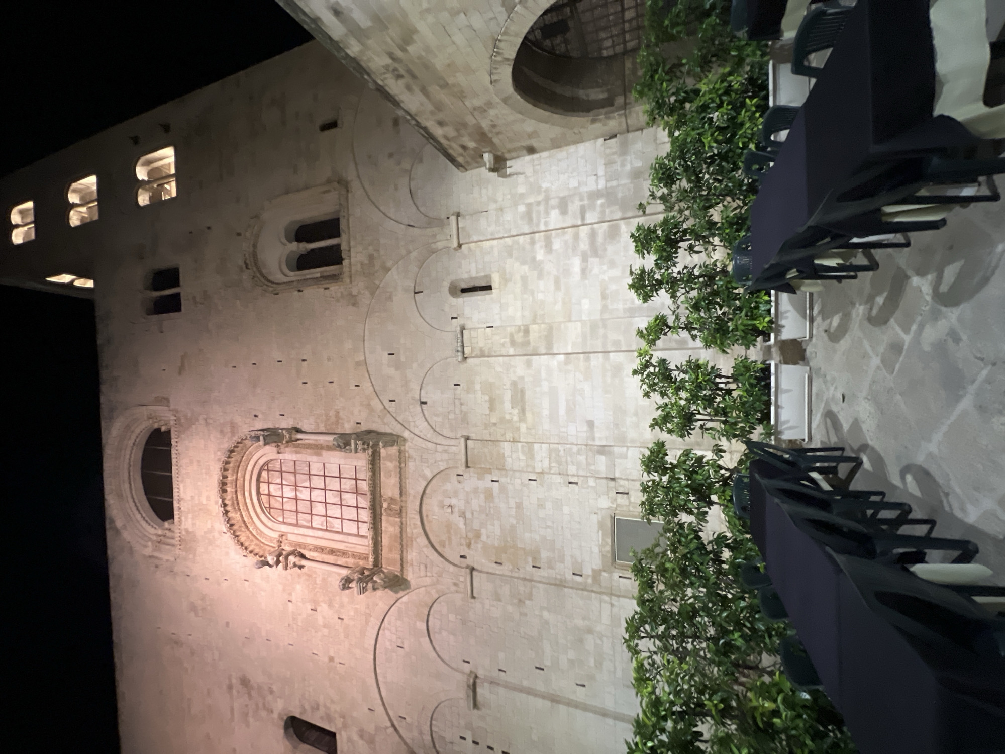 This image depicts an outdoor seating area set up at night in front of the Church of San Rocco in Bitonto, Italy. Two long tables, covered with black tablecloths and surrounded by dark plastic chairs, are positioned on a paved courtyard. Behind the tables, a row of green potted plants in white rectangular planters runs along a stone wall. The wall is part of the historic church, characterized by its stone facade, arched windows, and a tall, illuminated bell tower. The lighting in the scene is a mix of artificial lights illuminating the church facade and the tower, and the ambient darkness of the night sky. There are no people visible in the image.