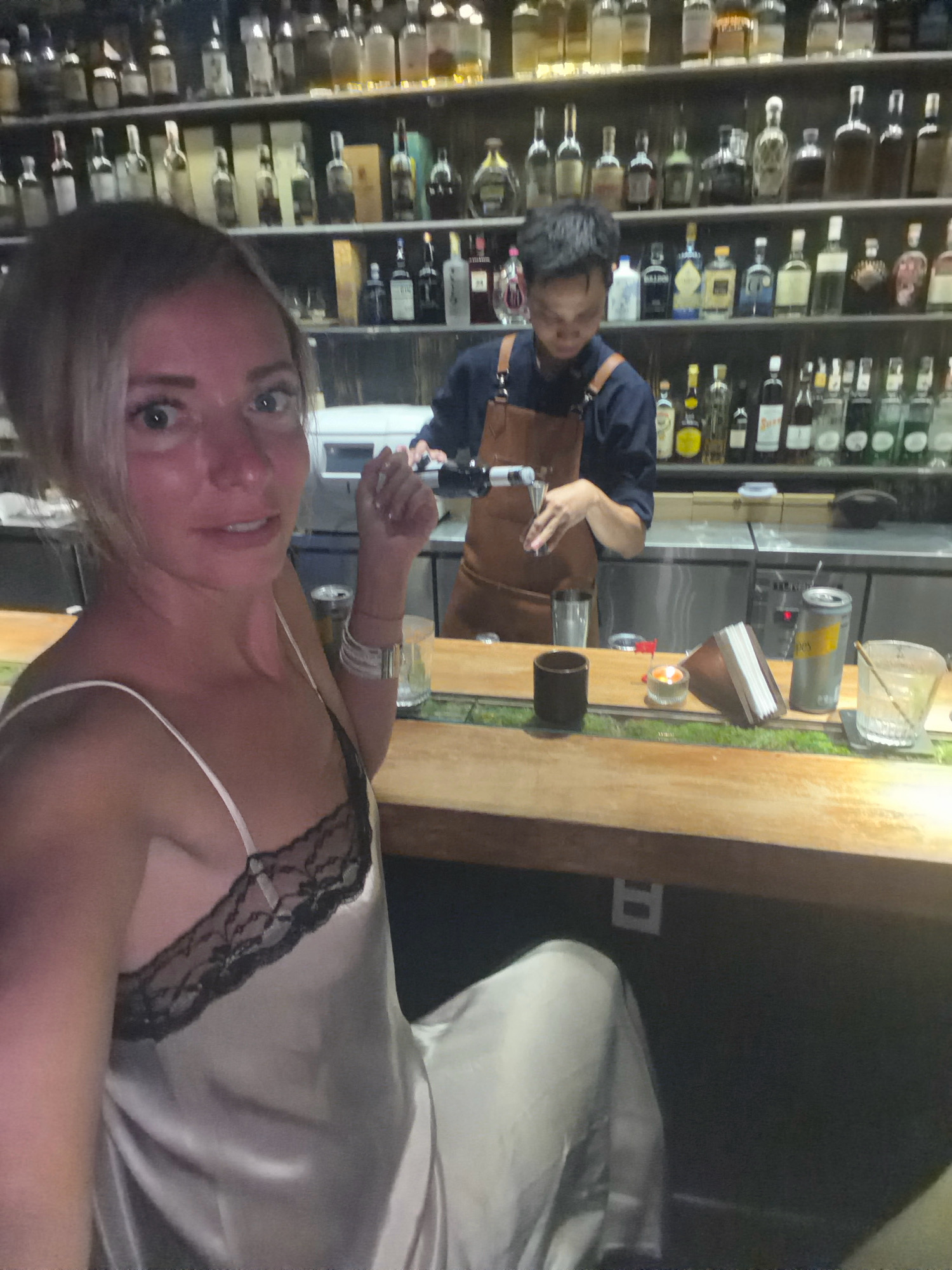 A young woman with blonde hair is taking a selfie at a bar. She is wearing a light-colored slip dress with black lace trim. Behind her, a bartender wearing an apron is preparing a drink. The bar is lined with shelves stocked with various bottles of liquor and glassware. A wooden counter separates the patrons from the bartender, and a variety of bar accessories, including glasses, a can, a napkin holder, and a small candle, are visible on the counter. The lighting is dim, suggesting an evening setting. The bar appears to be a sophisticated establishment, possibly a cocktail bar, with a wide selection of beverages displayed on the shelves. It is unknown if this scene is located in Da Lat, Vietnam.