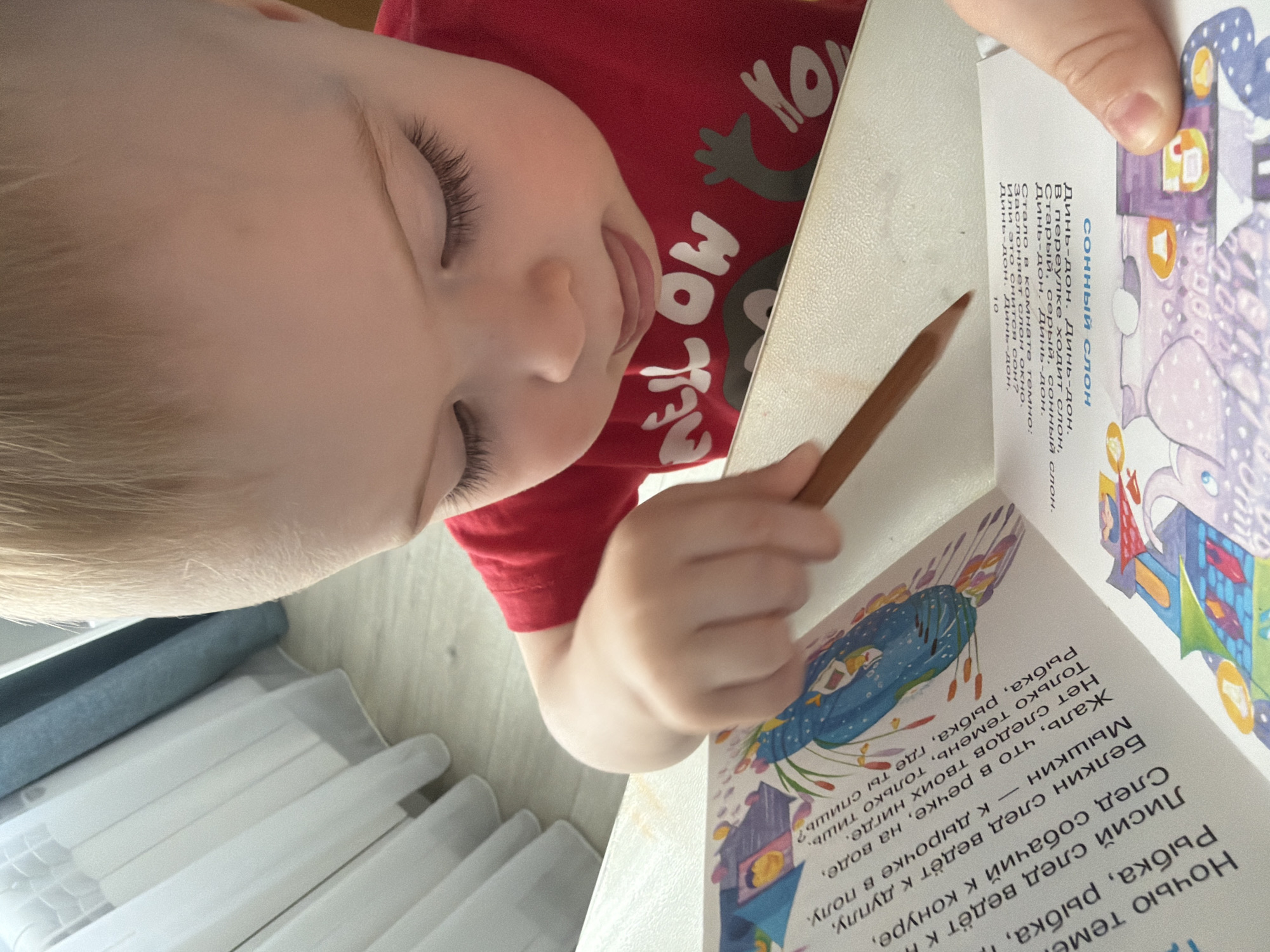 A young child in a red t-shirt is examining a book with illustrations and Russian text while holding a brown pencil. The book appears to be lying on a table, with the child's face and hands close to it. A set of white crayons in clear plastic packaging are visible in the lower left corner of the image.