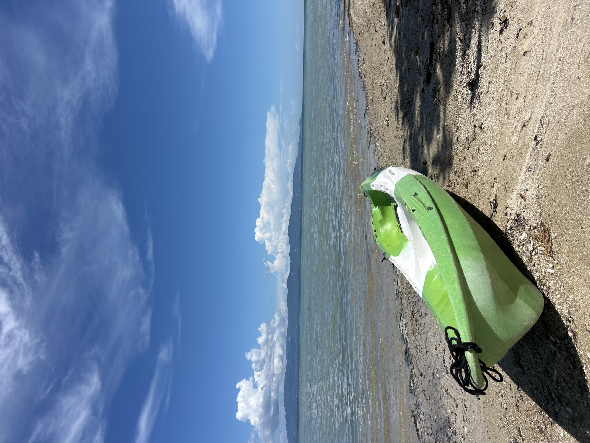 A bright green and white kayak rests on a sandy beach beside calm, light blue water under a bright blue sky filled with scattered white clouds. In the distance, a hazy mountain range stretches across the horizon. The foreground of the image shows the textured sand with some dried leaves and debris scattered on it, and shadows cast from overhead foliage.