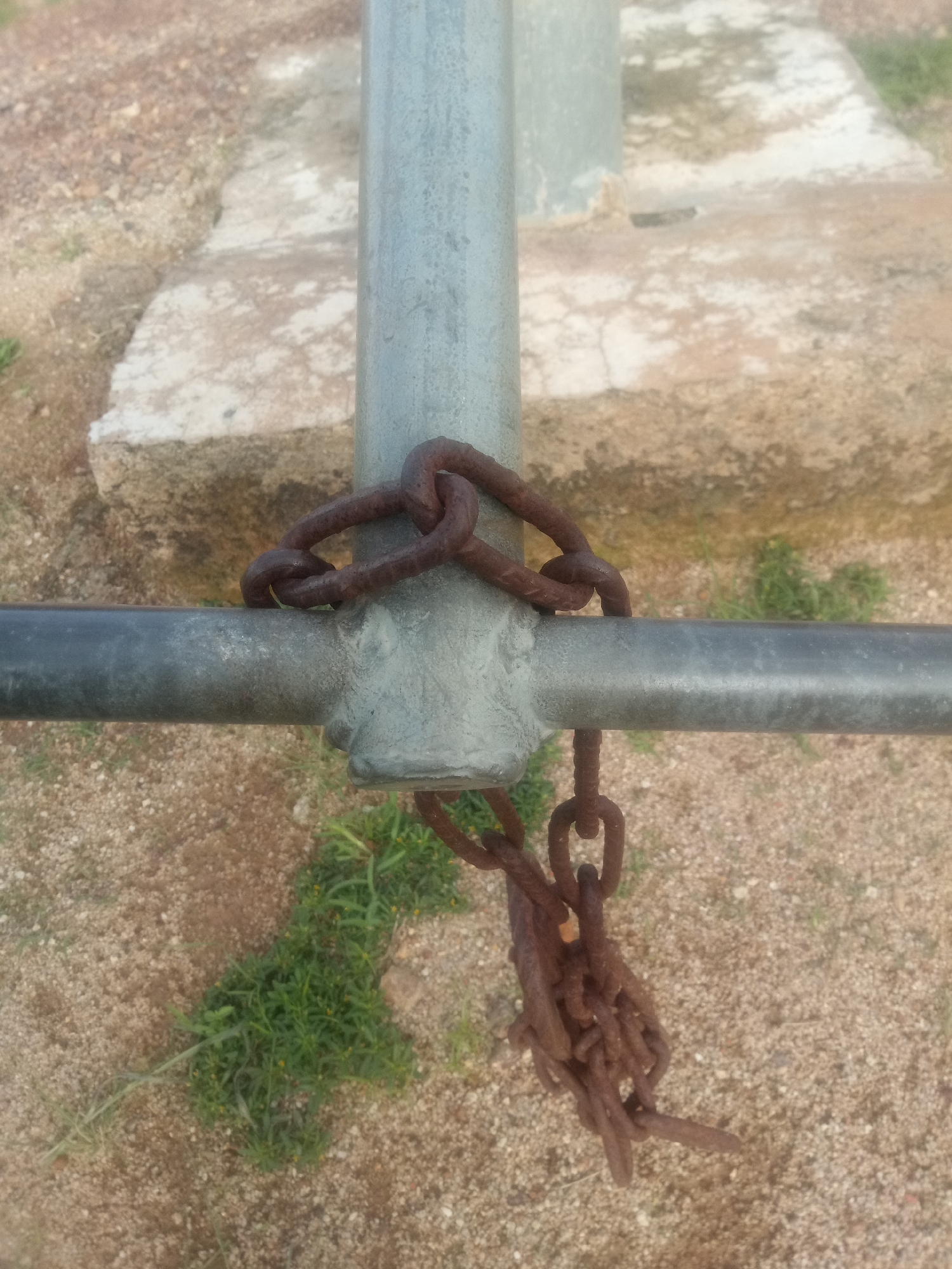 The image shows a close-up of a metal pole with a horizontal bar attached to it. Two rusty chains are wrapped around the pole and the bar. One chain is looped around the pole and the bar, while the other chain hangs down from the bar, forming a tangled cluster of links. The background consists of gravel and some scattered green vegetation. The scene appears to be outdoors, in an unspecified urban location. There are no people visible in the image.