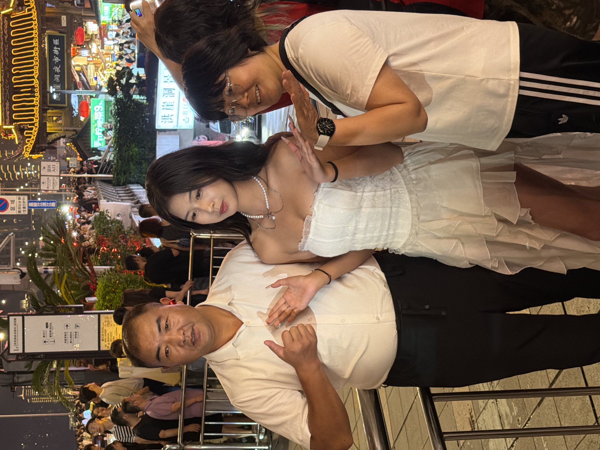This media file captures a lively scene at night in Yuzhong District, China, likely near the popular Hongyadong area. The foreground features three individuals. Closest to the viewer, an Asian man with short hair and a light-colored polo shirt is smiling and looking towards the camera, with his right hand making a "shaka" gesture. Behind him and slightly to his left, a young Asian woman with long dark hair is wearing a white strapless dress adorned with pearls. She is looking downwards with a neutral expression. To her left and slightly behind her, an Asian woman with dark hair and glasses is looking at the camera and smiling, with her hand raised as if to wave or block the sun.
The background is a bustling street scene at night, illuminated by numerous colorful lights and neon signs. The text "朝天门" and "崖洞夜市街區" (Cliffside Night Market Street Area) are visible on signs, indicating the setting. There are also signs with Chinese characters and directional arrows, suggesting a tourist or commercial area. The overall atmosphere appears festive and crowded, with many people visible in the background, though they are less distinct. The time of day is clearly night, indicated by the artificial lighting and the general ambiance. The weather is not explicitly discernible but the clothing suggests a mild evening.
